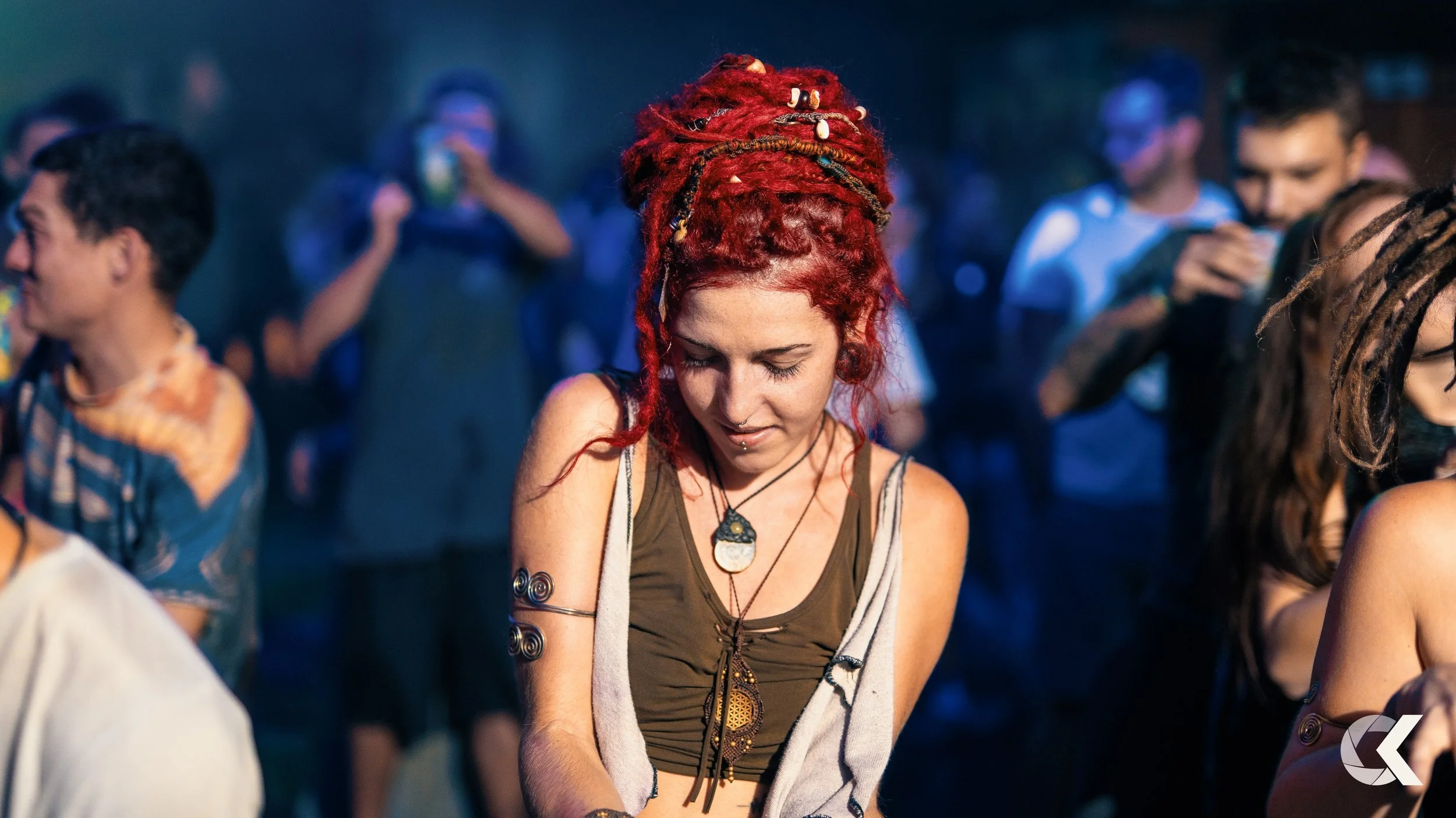 A woman with red dreadlocks, wearing multiple necklaces, appears to be enjoying a music festival or outdoor event at night, surrounded by other people.