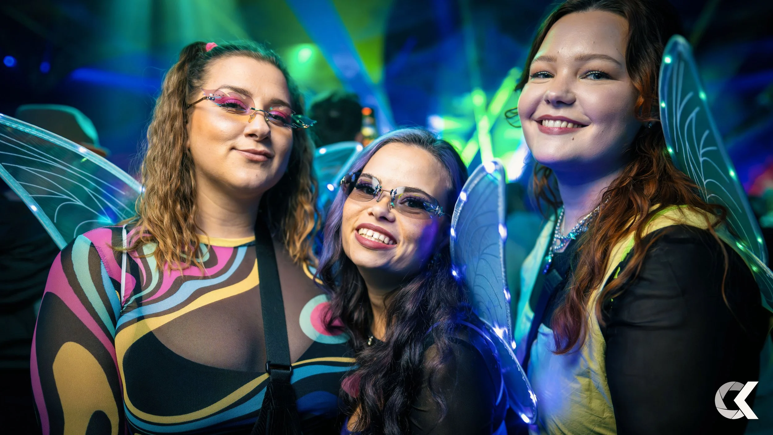 Three women dressed as fairies at a costume party with colorful lighting and fairy wings.