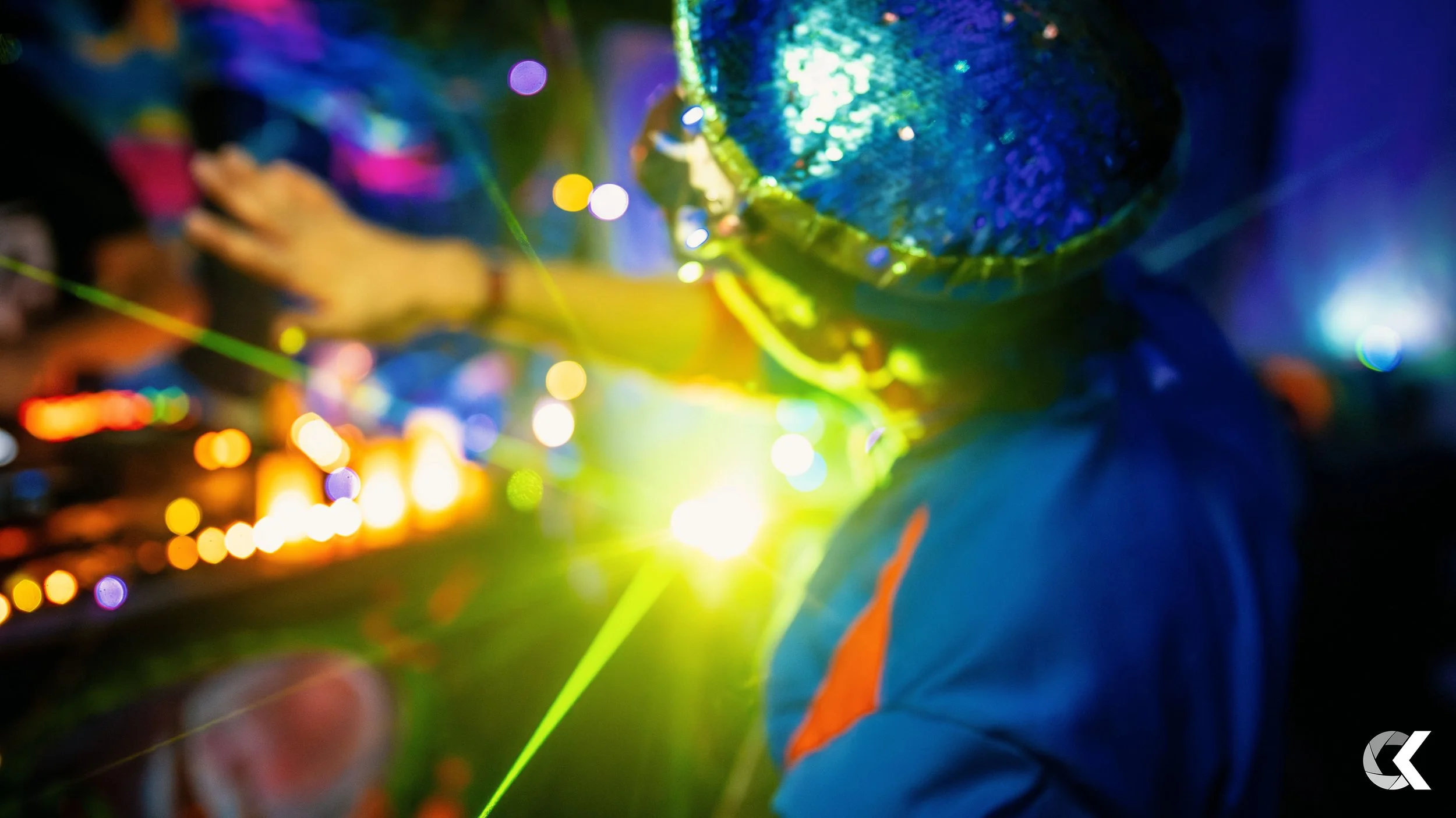 A person wearing a shiny blue hat and a blue jacket is at a festive event with colorful lights and laser effects in the background.