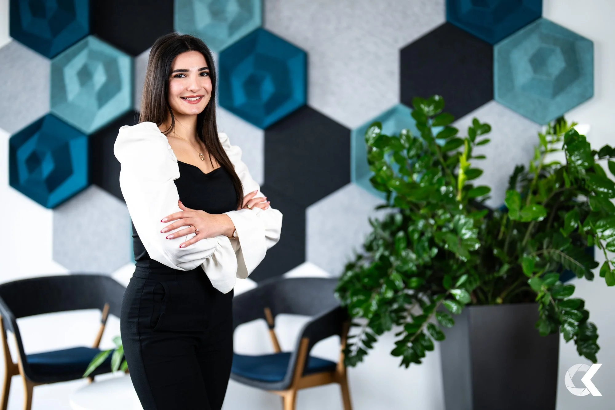 A woman standing with arms crossed in a modern office space, smiling, with a patterned wall and chairs in the background, and a large green plant in the foreground.