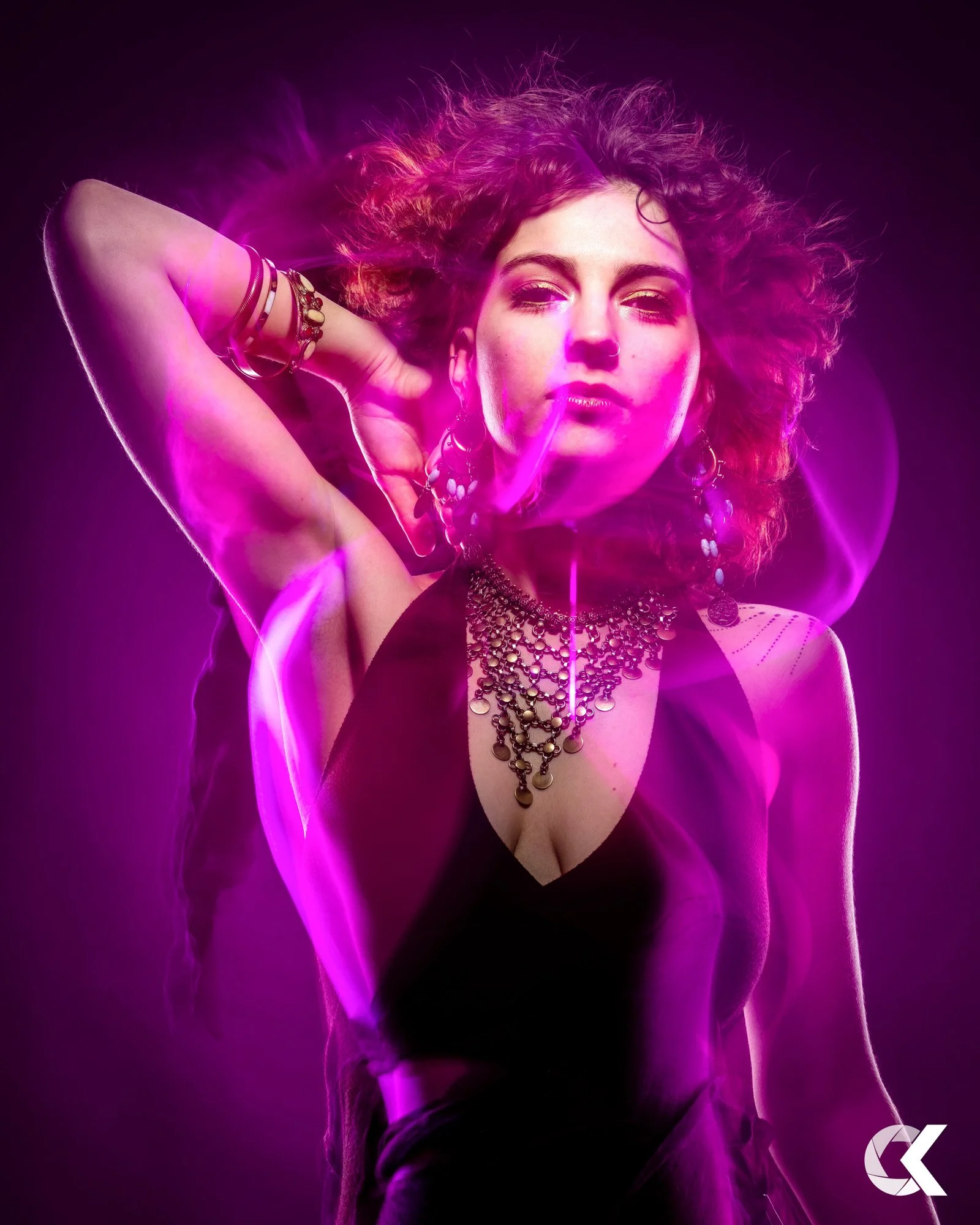 Junge Frau mit vielen Schmuckstücken, dunklem Kleid und surrealem, violettem Licht, die einen relaxed Ausdruck zeigt.