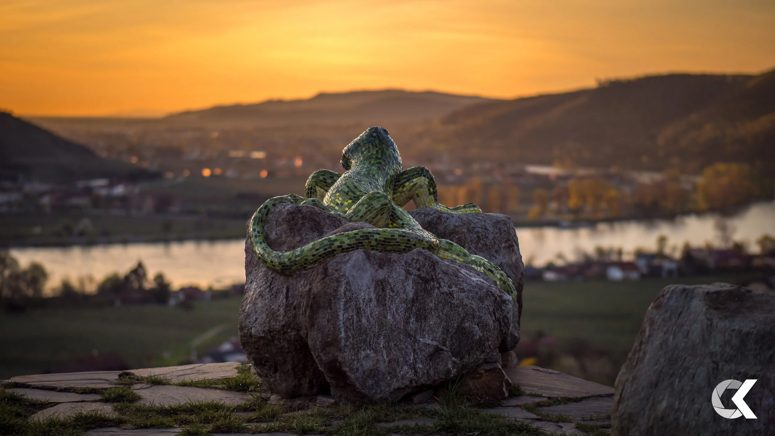 A decorative sculpture of a lizard with green mosaic tiles sitting on a rock during sunset, overlooking a landscape with water, houses, and hills.