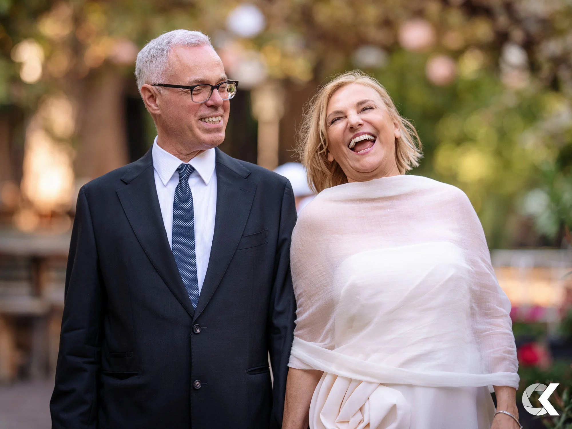 A smiling woman wearing a white dress and a shawl, walking beside a man in a black suit, both outdoors with a blurred background of trees and pink blossoms.