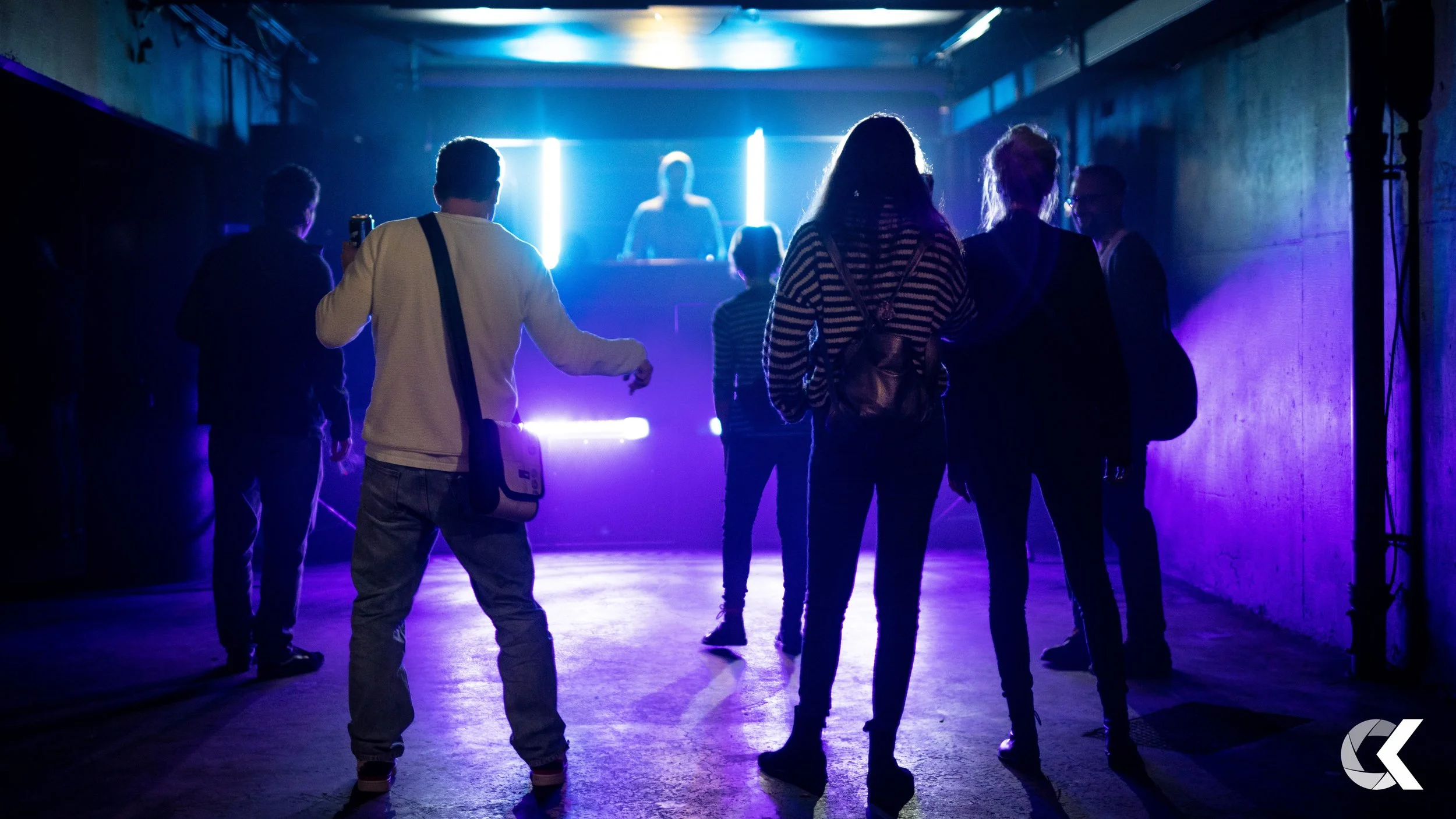 People dancing at a nightclub with blue and purple neon lights, DJ on stage in background.