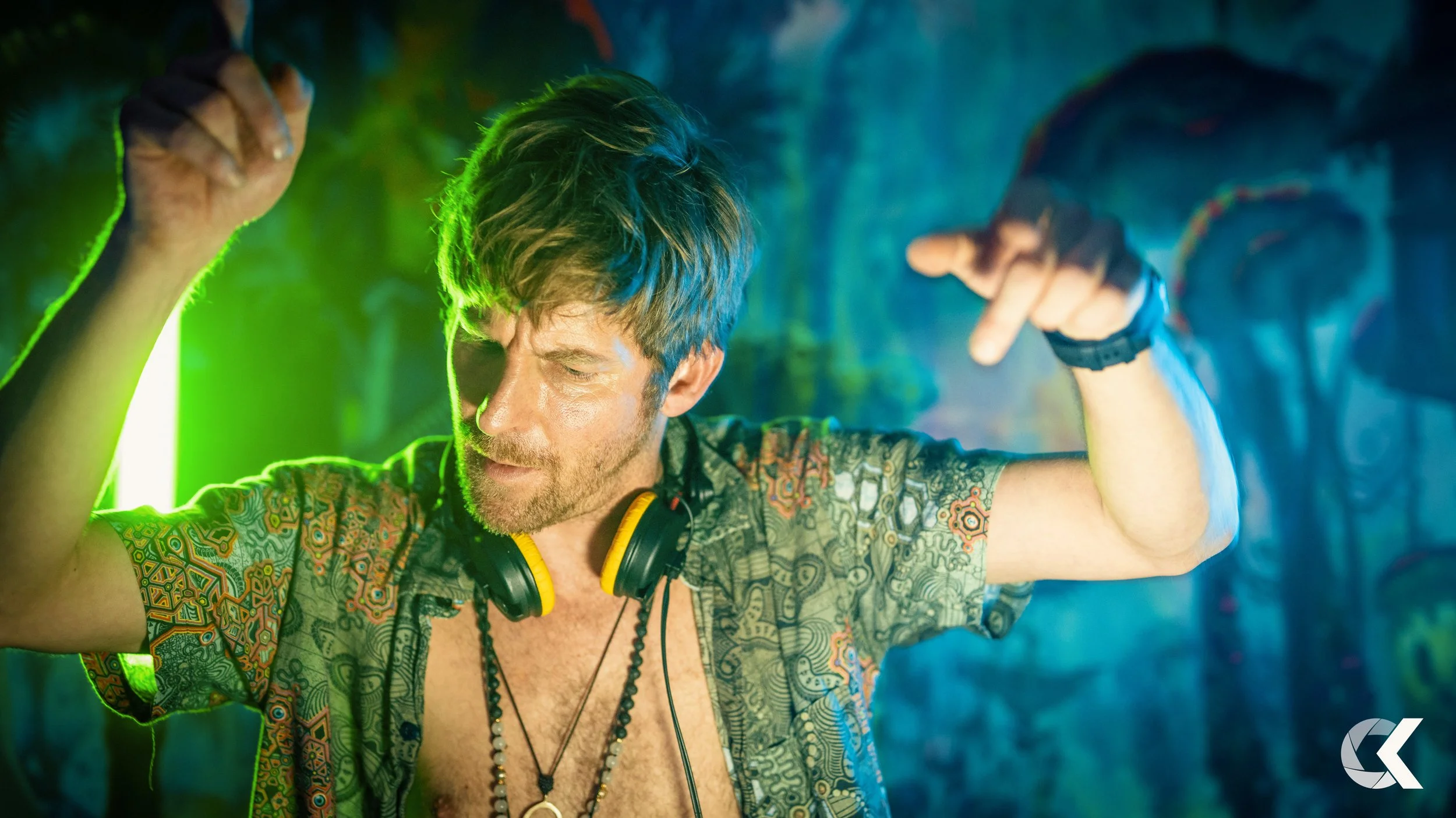 A young man with brown hair and a beard, wearing headphones around his neck and a colorful patterned shirt, is dancing with his eyes closed in a dimly lit room with green and blue lighting.