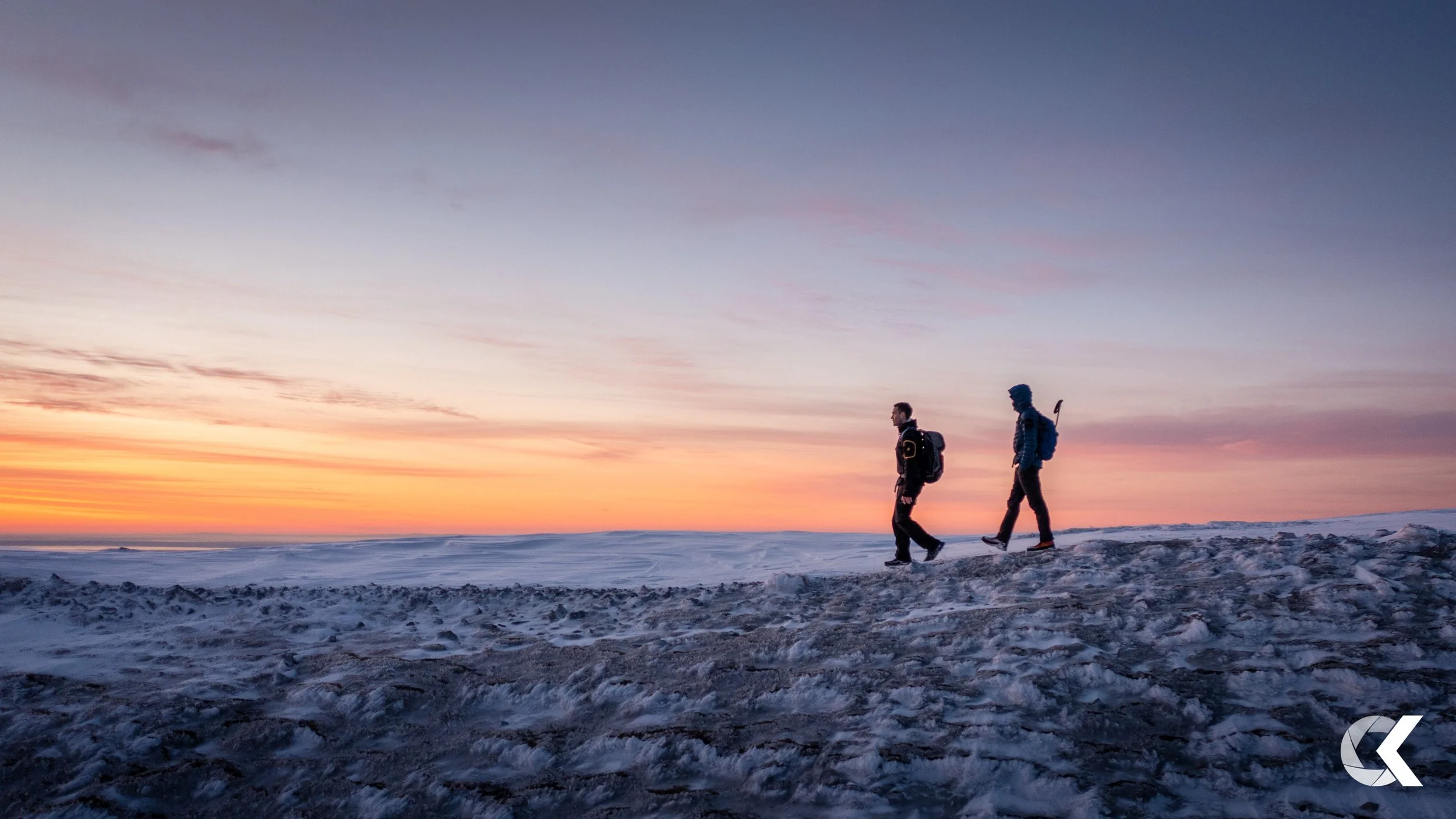 Two hikers walking on snow-covered terrain during sunset with a colorful sky in the background.