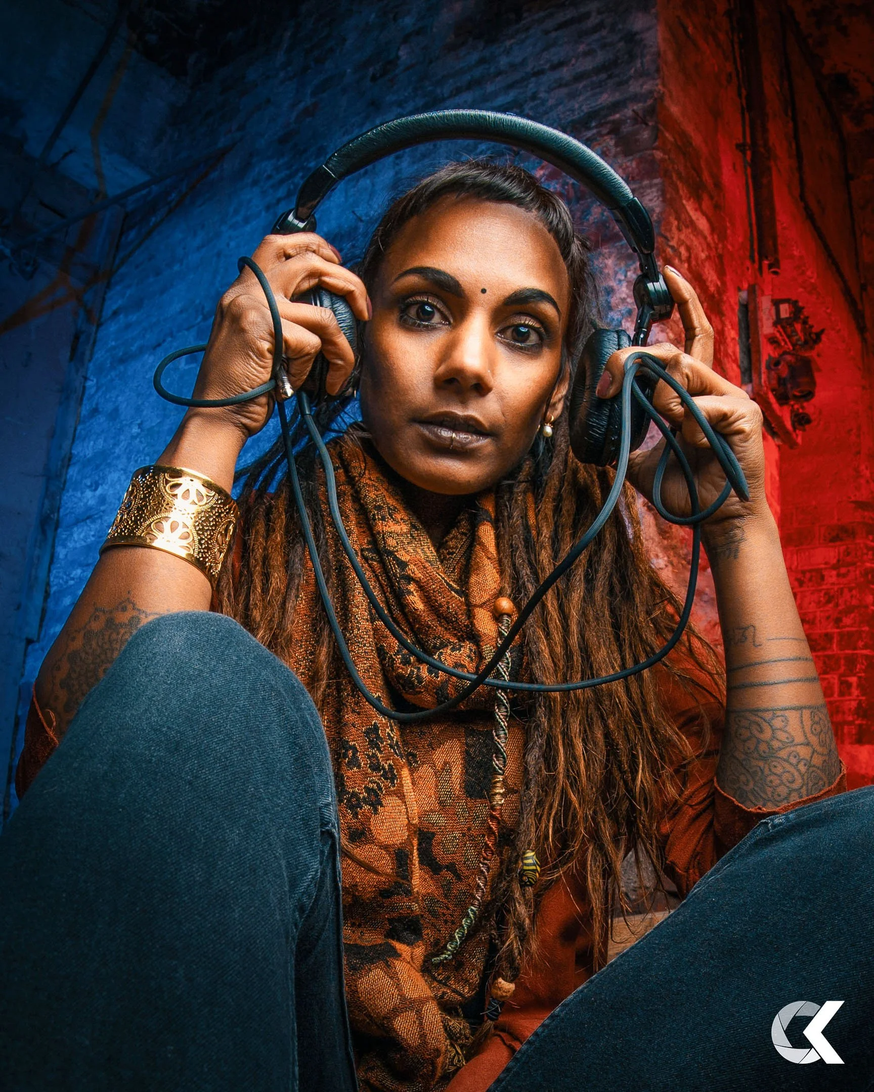 A woman with dreadlocks and tattoos poses holding a headset, with colorful blue and red lighting in the background.