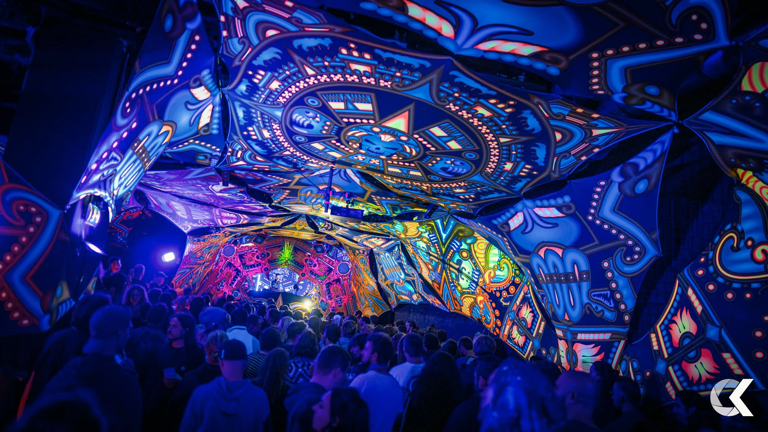 A large crowd of people at a vibrant, colorful light art installation with intricate, neon patterns projected on a tunnel-like structure.