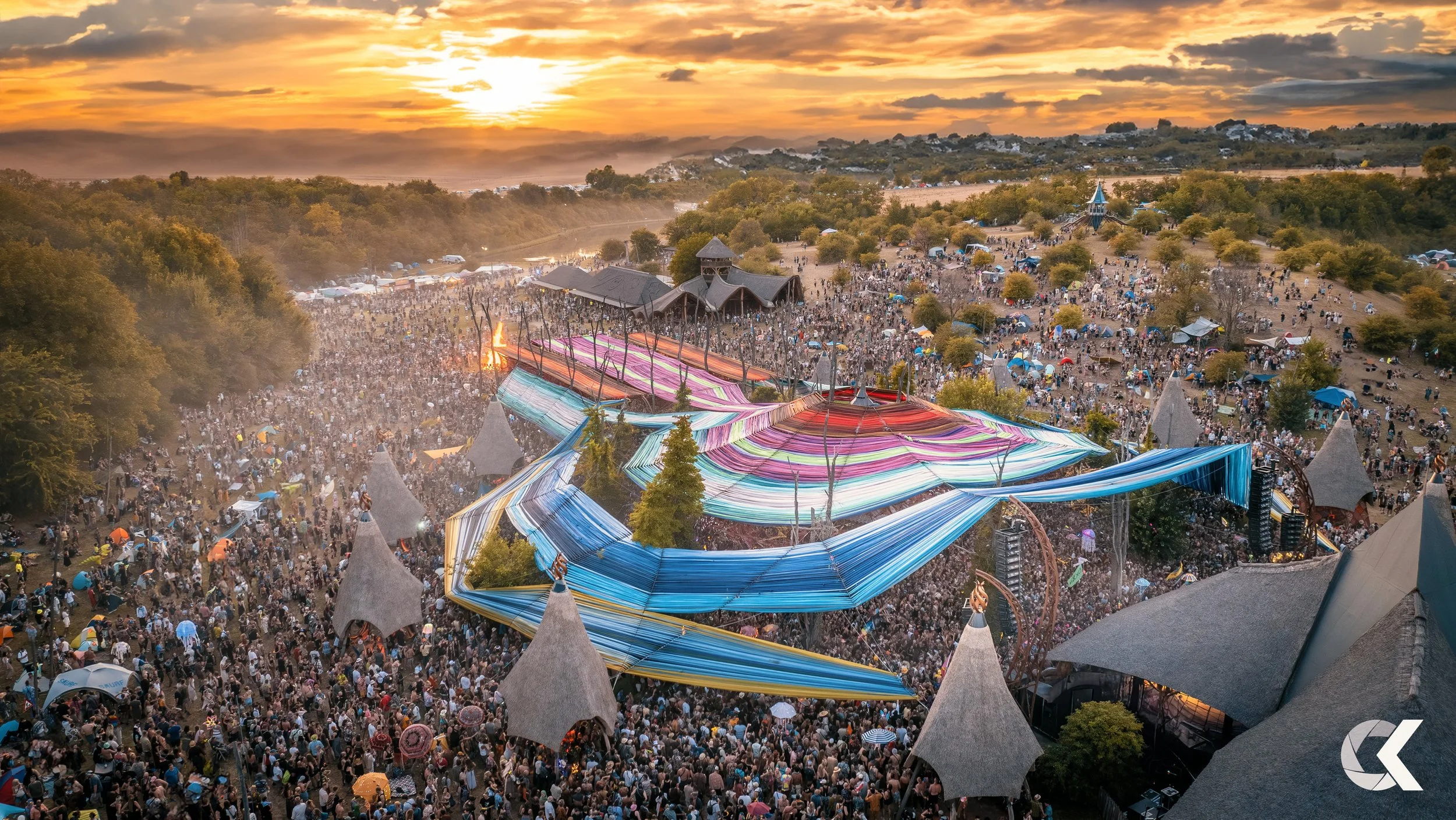 A large outdoor music festival at sunset with a crowd of people, colorful fabric canopies, trees, and festival structures visible.
