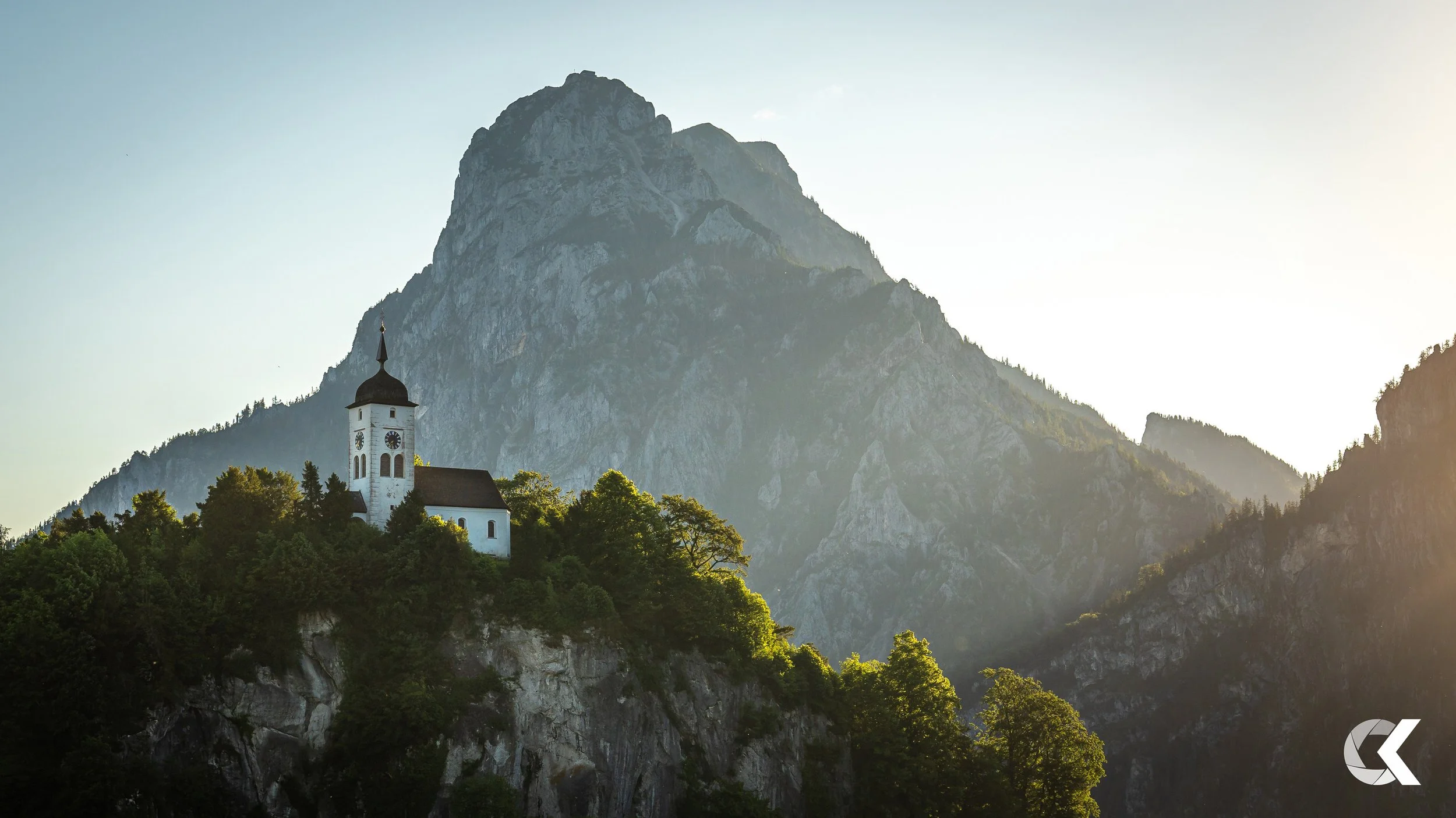 A white church with a black dome and clock tower on a lush green hillside at the foot of a large mountain range during sunset.