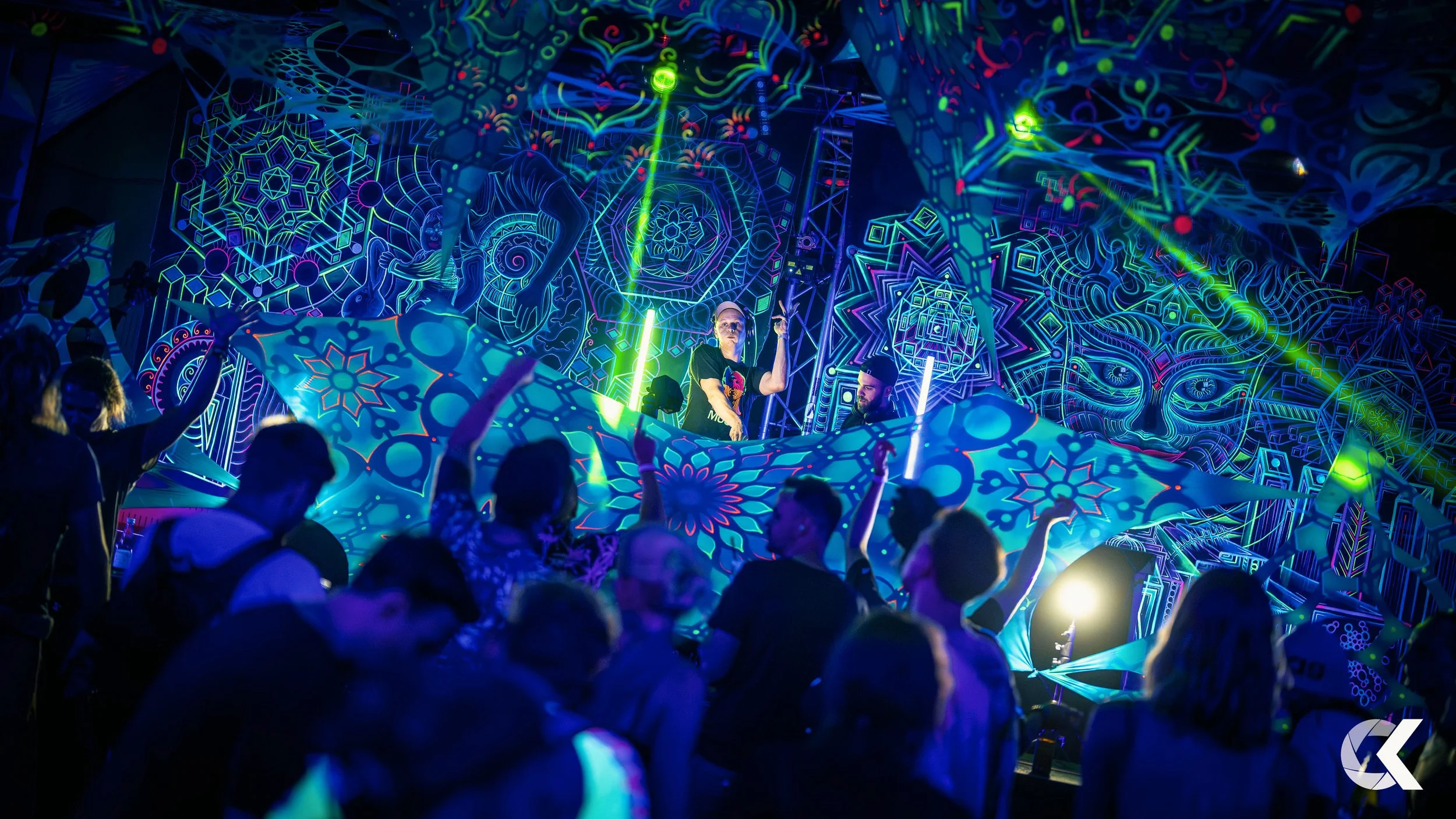 A vibrant electronic dance music concert with neon geometric and psychedelic patterns on the backdrop and LED lighting, featuring a DJ performing on stage with a crowd dancing in front.