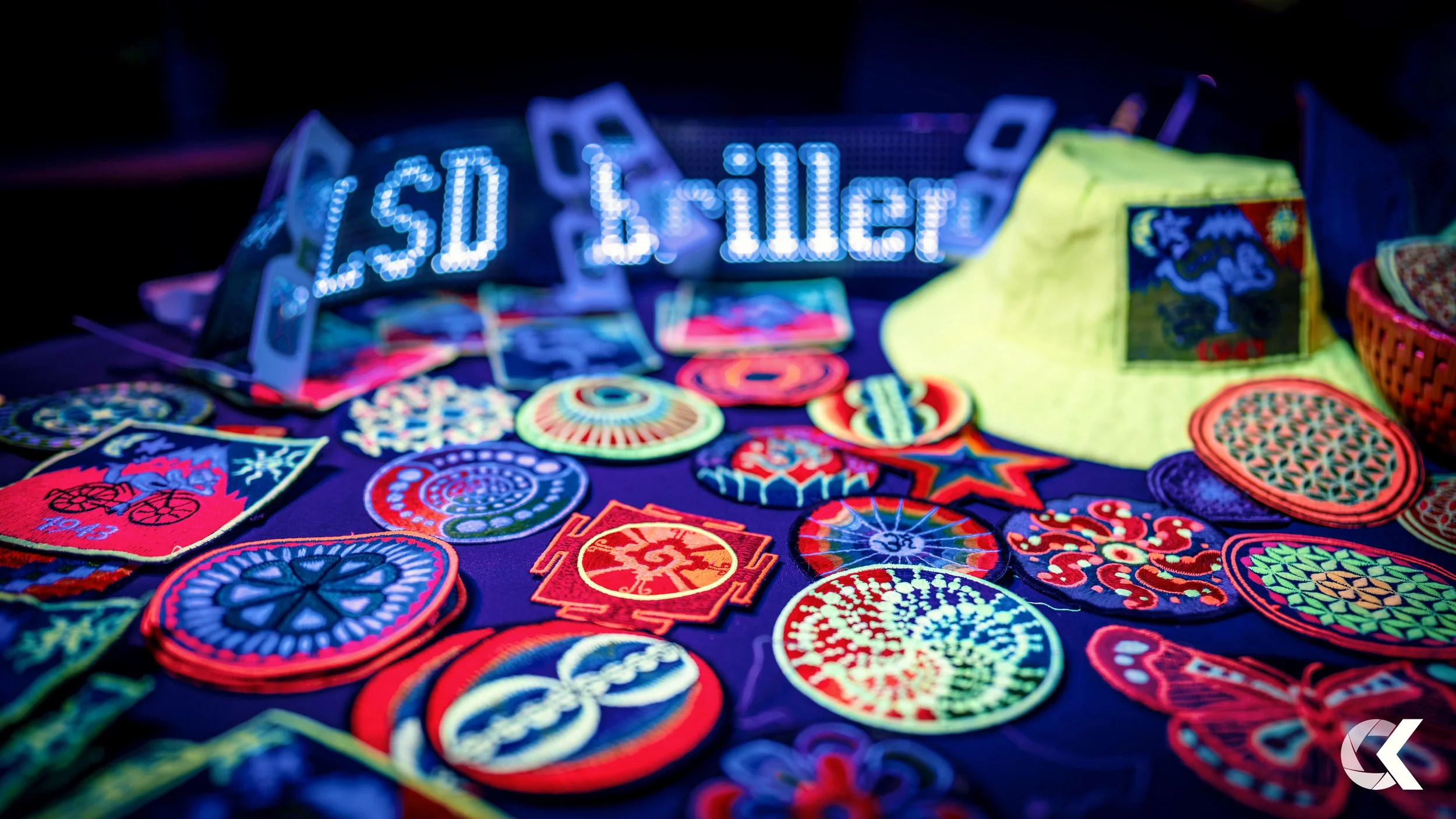 A table with embroidered patches, a neon sign that reads '$1.00 Seller,' and a lime green cap with a logo, illuminated by vibrant blacklight.