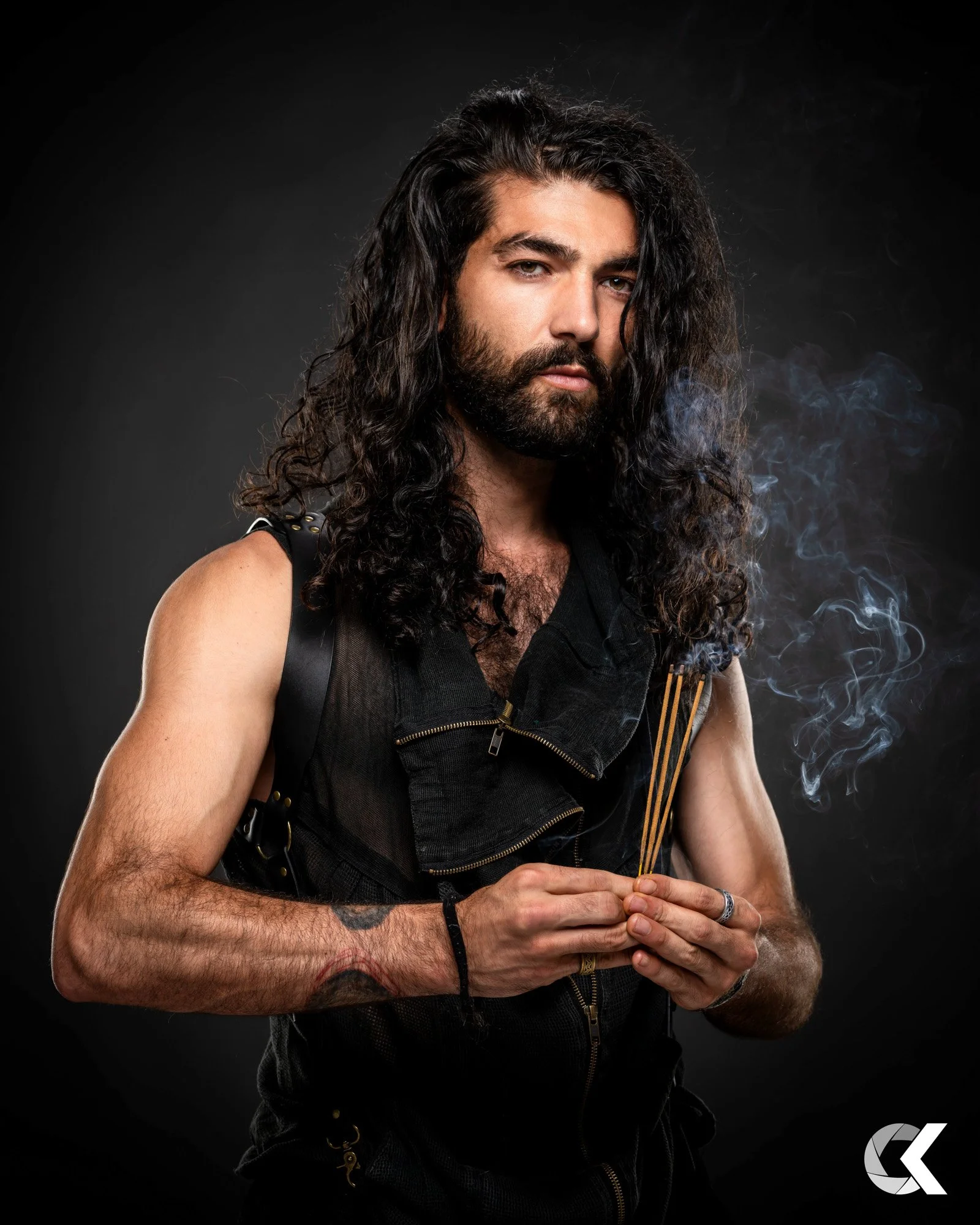 Junge Mann mit langem, dunklem, lockigem Haar und Bart, trägt schwarze Kleidung, hält brennende Yucca-Stäbchen, schwarze Hintergrund.