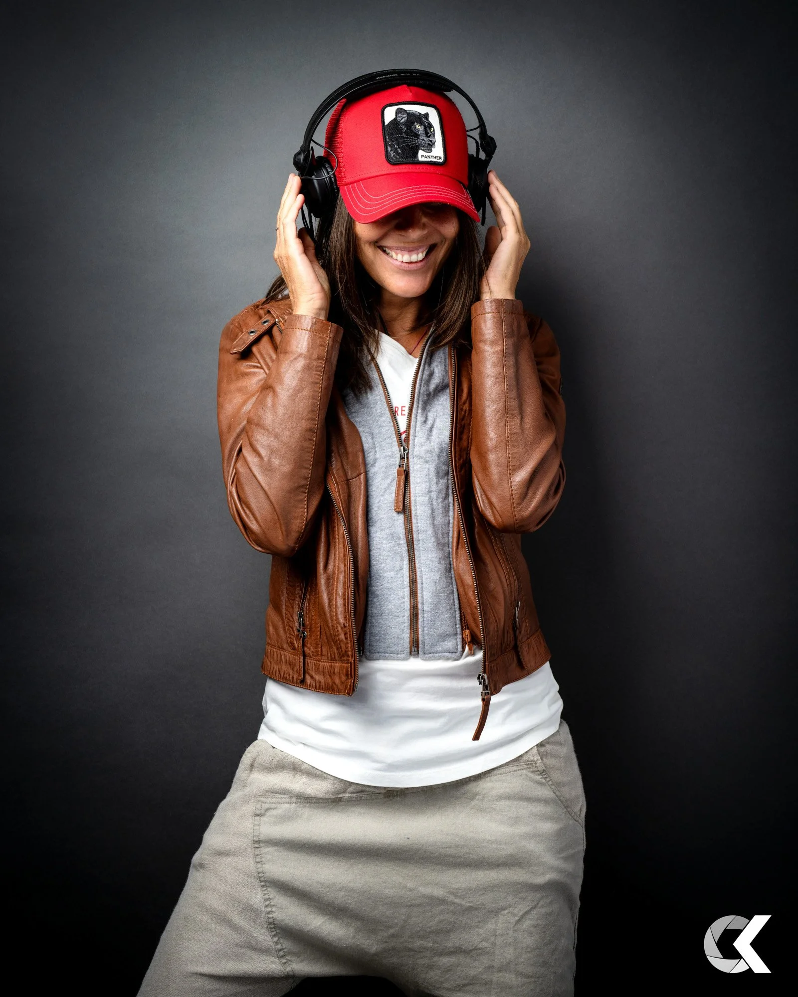 Woman wearing a red baseball cap with panther patch, brown leather jacket, white T-shirt, and gray pants, smiling and holding headphones on her head against gray background.