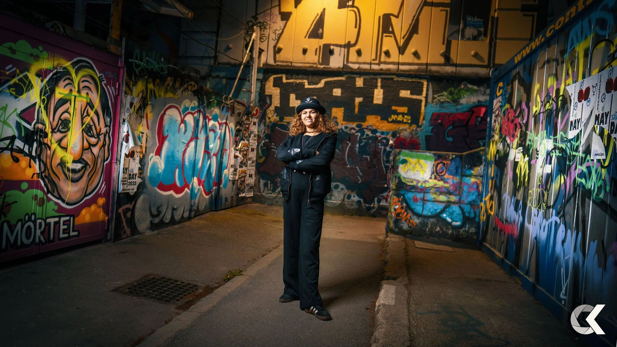 A woman standing with arms crossed in a graffiti-covered alleyway at night, wearing a black jacket, black pants, and a black hat, surrounded by colorful street art.