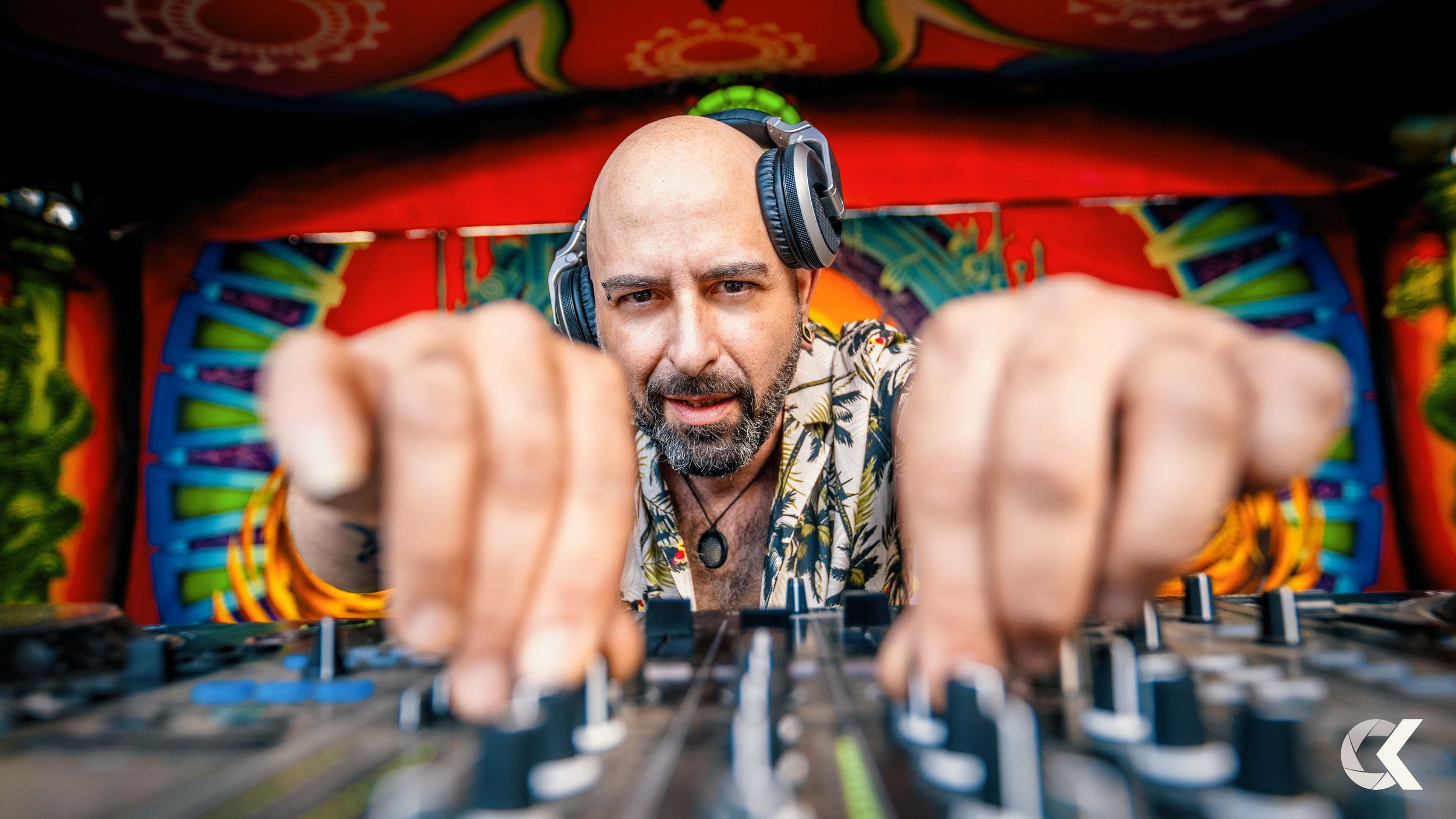 DJ wearing headphones, looking into camera, adjusting turntables at a vibrant, colorful party or festival booth.