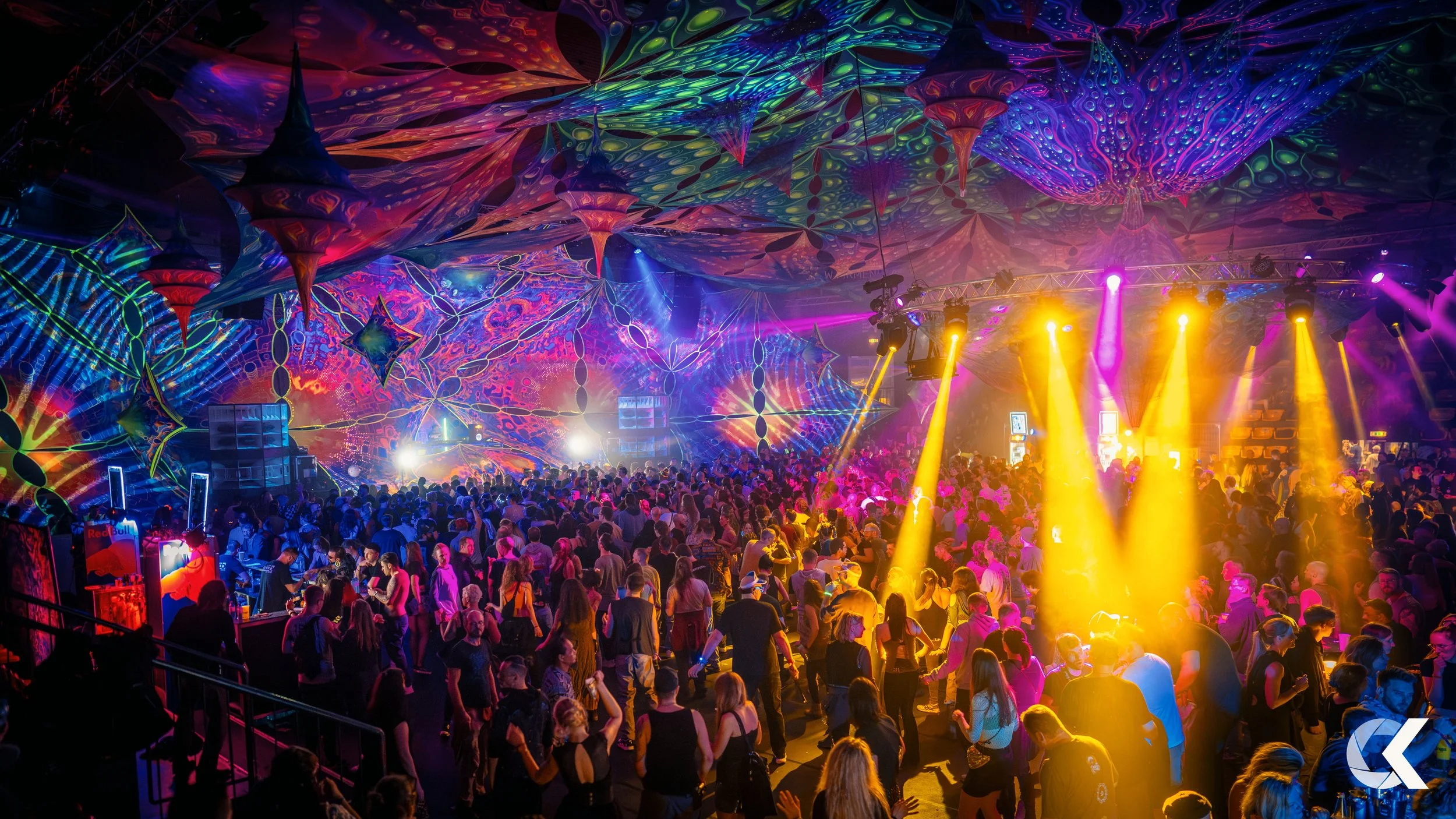 A vibrant indoor dance party with colorful psychedelic decorations and neon lighting, filled with a large crowd of people dancing and socializing under a decorated ceiling.