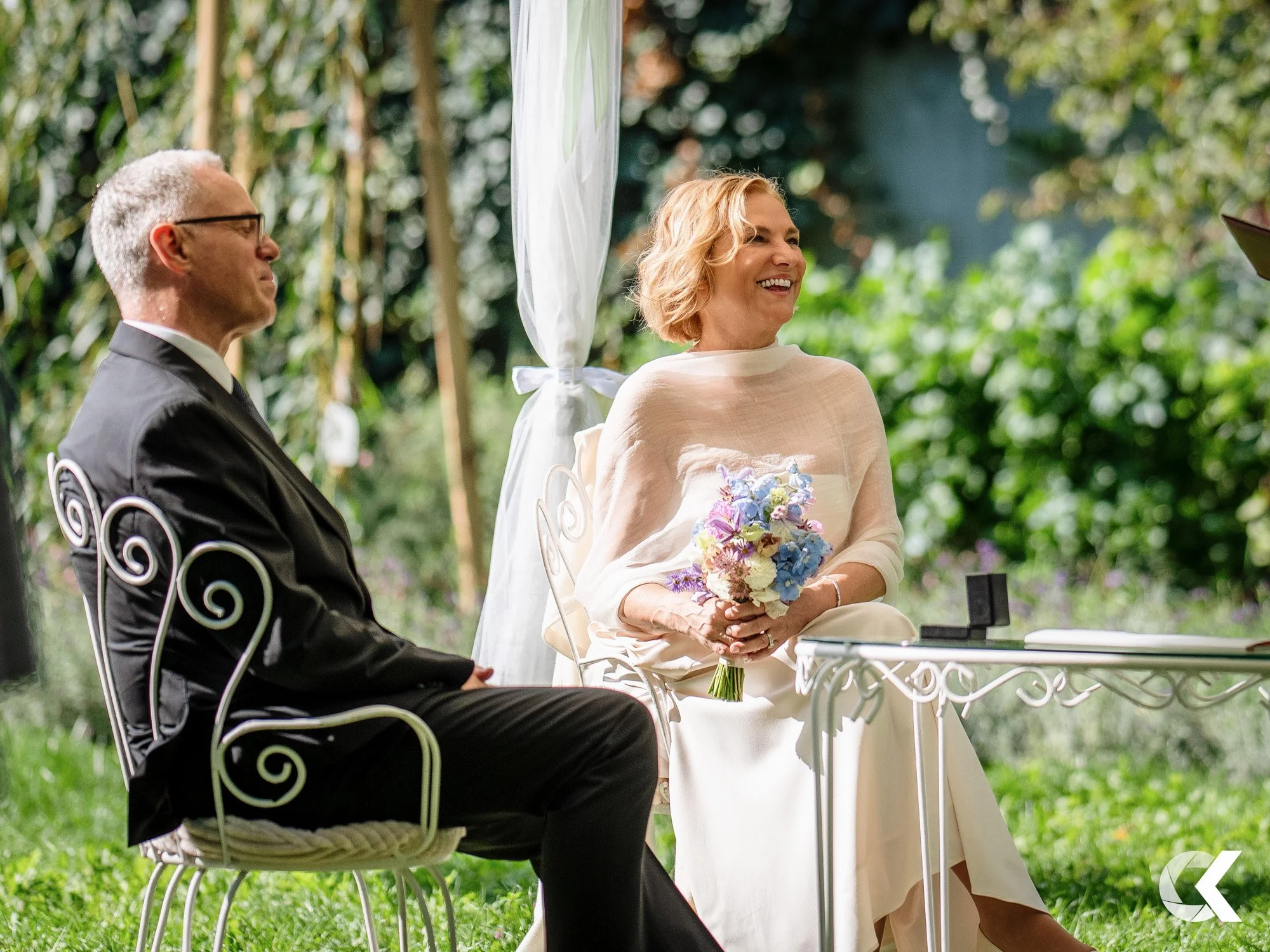 A woman in a cream dress holding a bouquet of purple, blue, white, and pink flowers, smiling during an outdoor ceremony with a man in a black suit and glasses seated beside her.