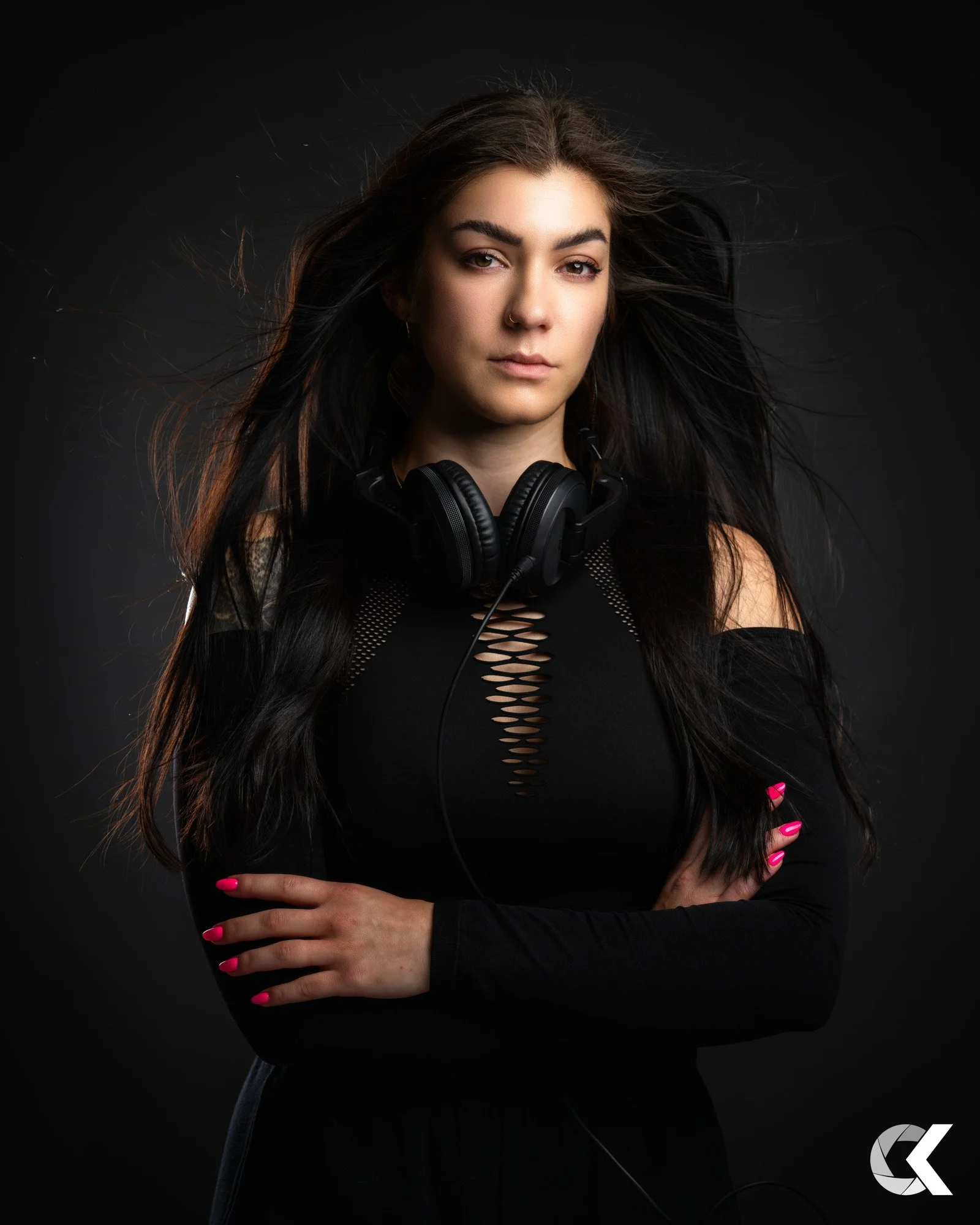 A young woman with long dark hair, light skin, and pink painted nails, poses with arms crossed, wearing a black top with cut-out details and headphones around her neck, against a dark background.
