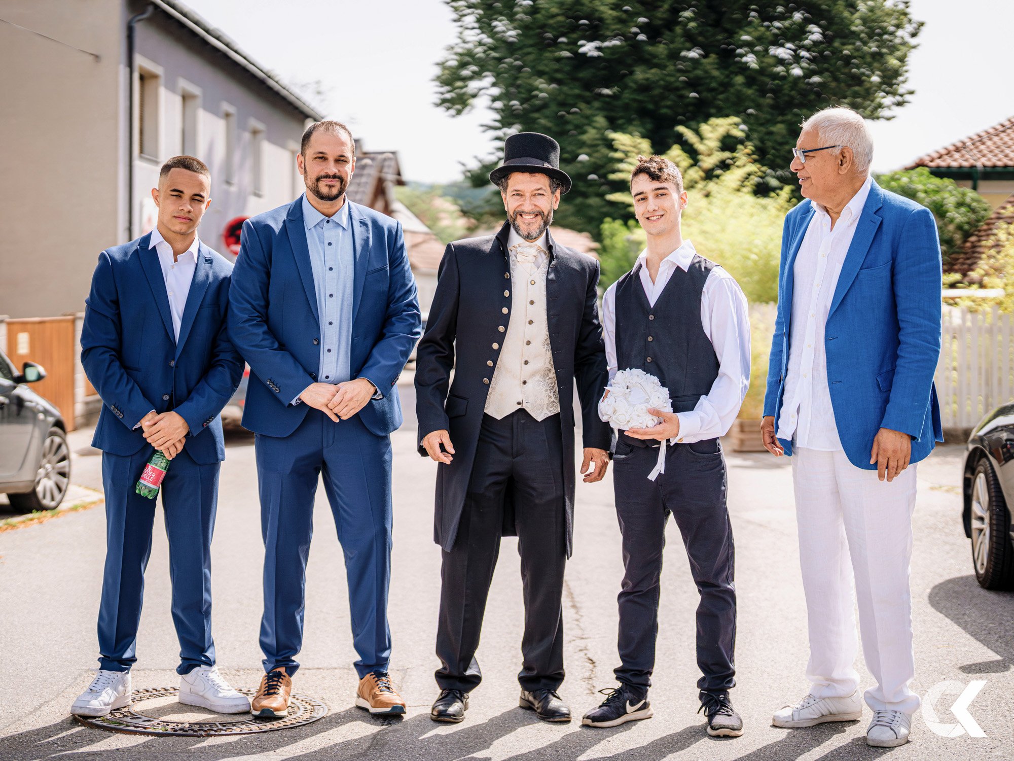 Group of men dressed in suits and formal attire standing outdoors in a residential neighborhood, with a young man holding a bouquet of white flowers.