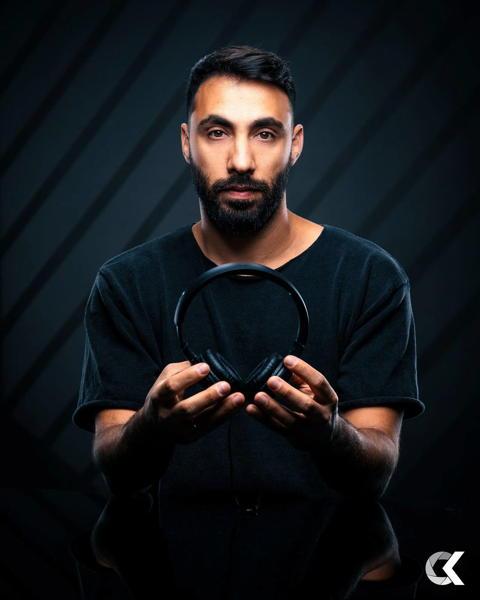 A bearded man in a black shirt holding black headphones with both hands, looking at the camera, against a dark background with diagonal lines.