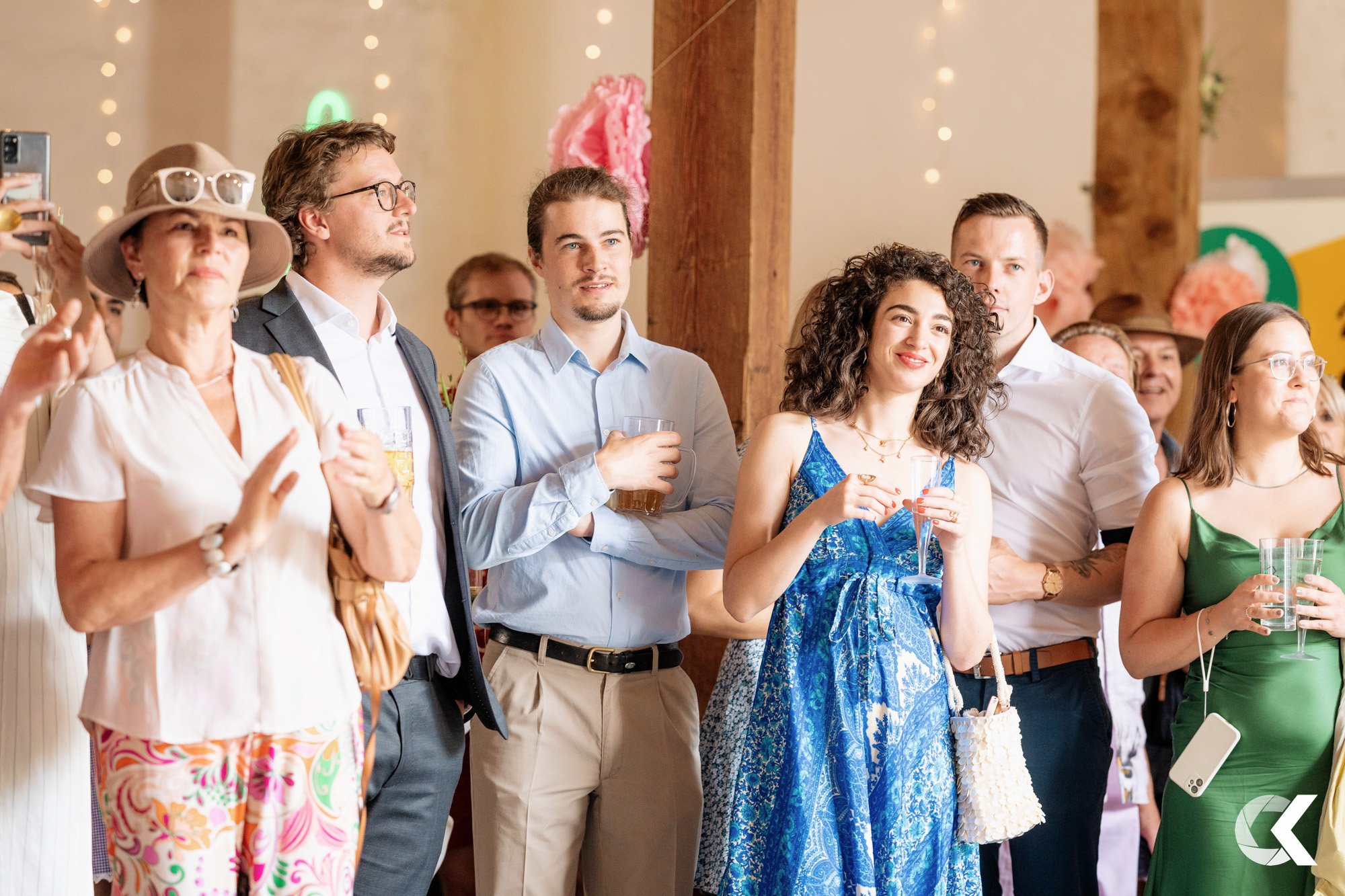 Group of people at a social gathering, some holding drinks, dressed in casual and semi-formal attire, indoor setting with decorative lights.