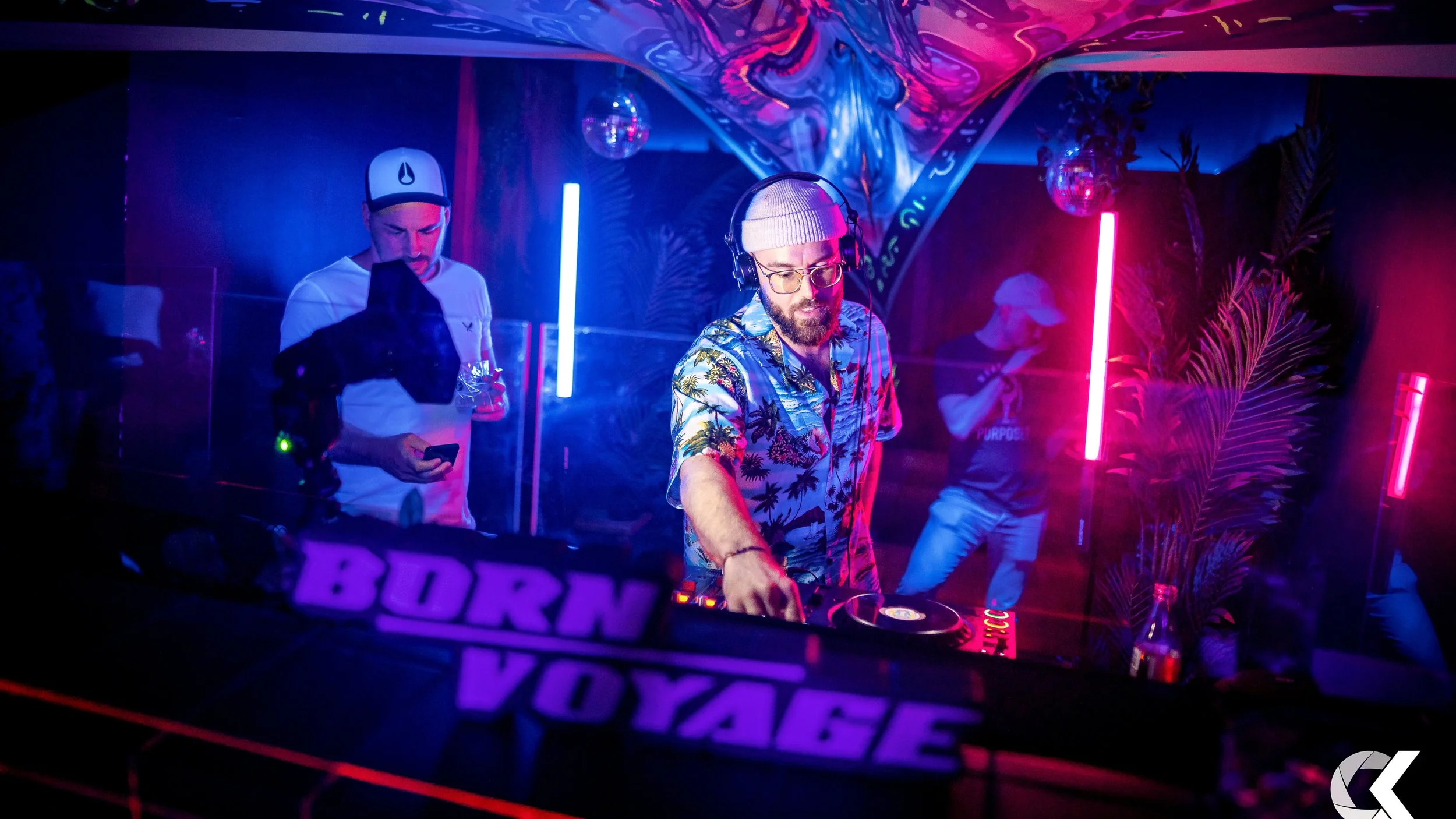 A DJ wearing glasses, a baseball cap, and a Hawaiian shirt is mixing music at a nightclub with neon lighting, plants, and a colorful art installation in the background.