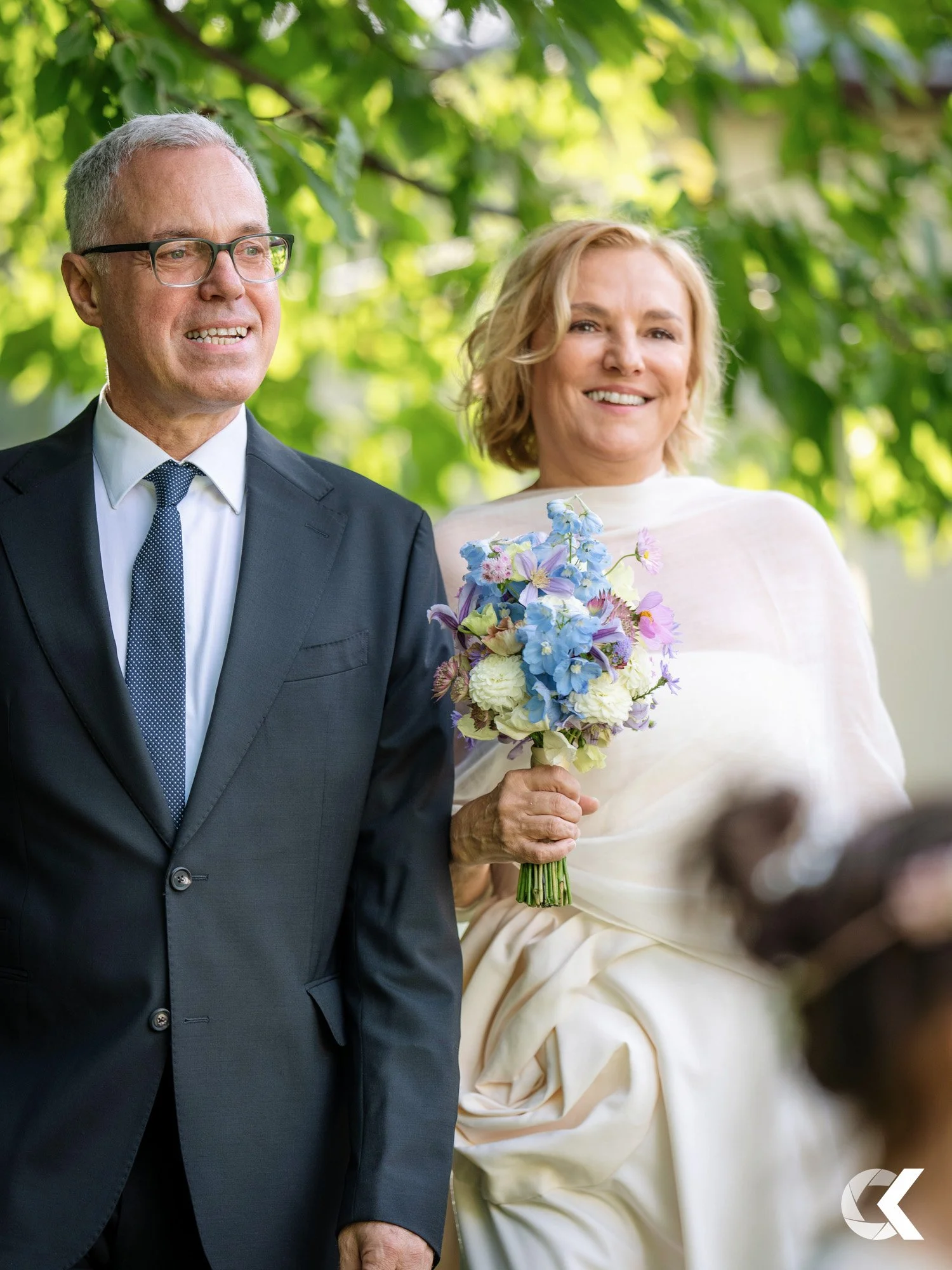 A middle-aged man in a black suit, white shirt, and blue tie, with glasses, standing next to a woman in a white dress holding a bouquet of purple, blue, and white flowers, outdoors under green leafy trees.