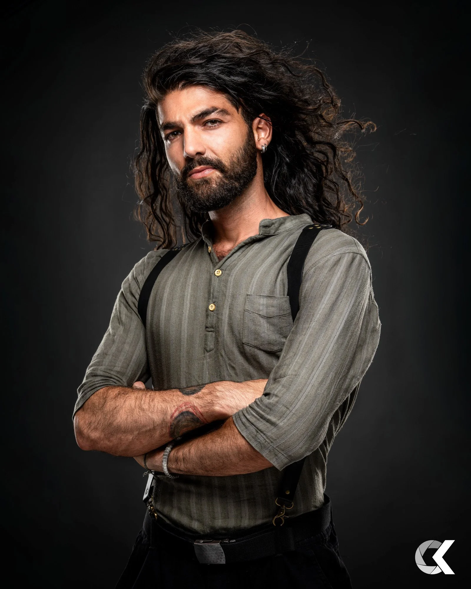 Porträt eines Mannes mit langem, lockigem Haar und Bart vor dunklem Hintergrund, trägt ein grünes Hemd mit aufgeknöpfter Kragenlinie, schwarze Hosenträger und Armbänder