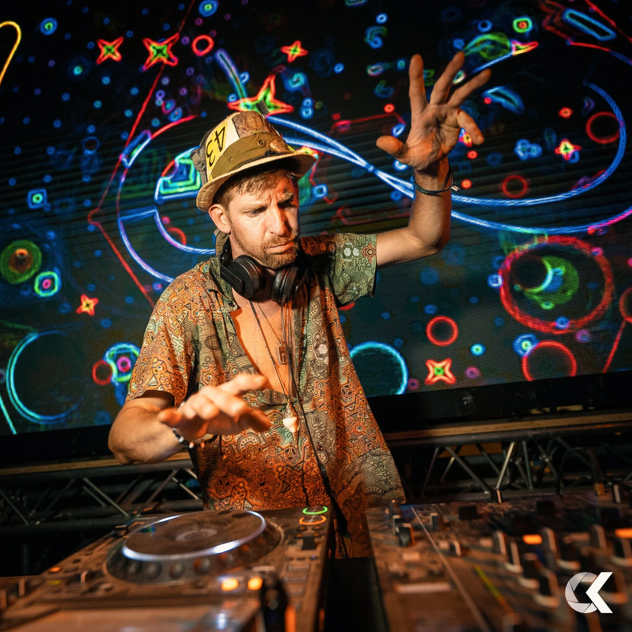 A DJ wearing a patterned shirt, headphones, and a hat is performing at a nightclub. The background features a digital display with colorful, abstract neon patterns and shapes, creating a lively and energetic atmosphere.