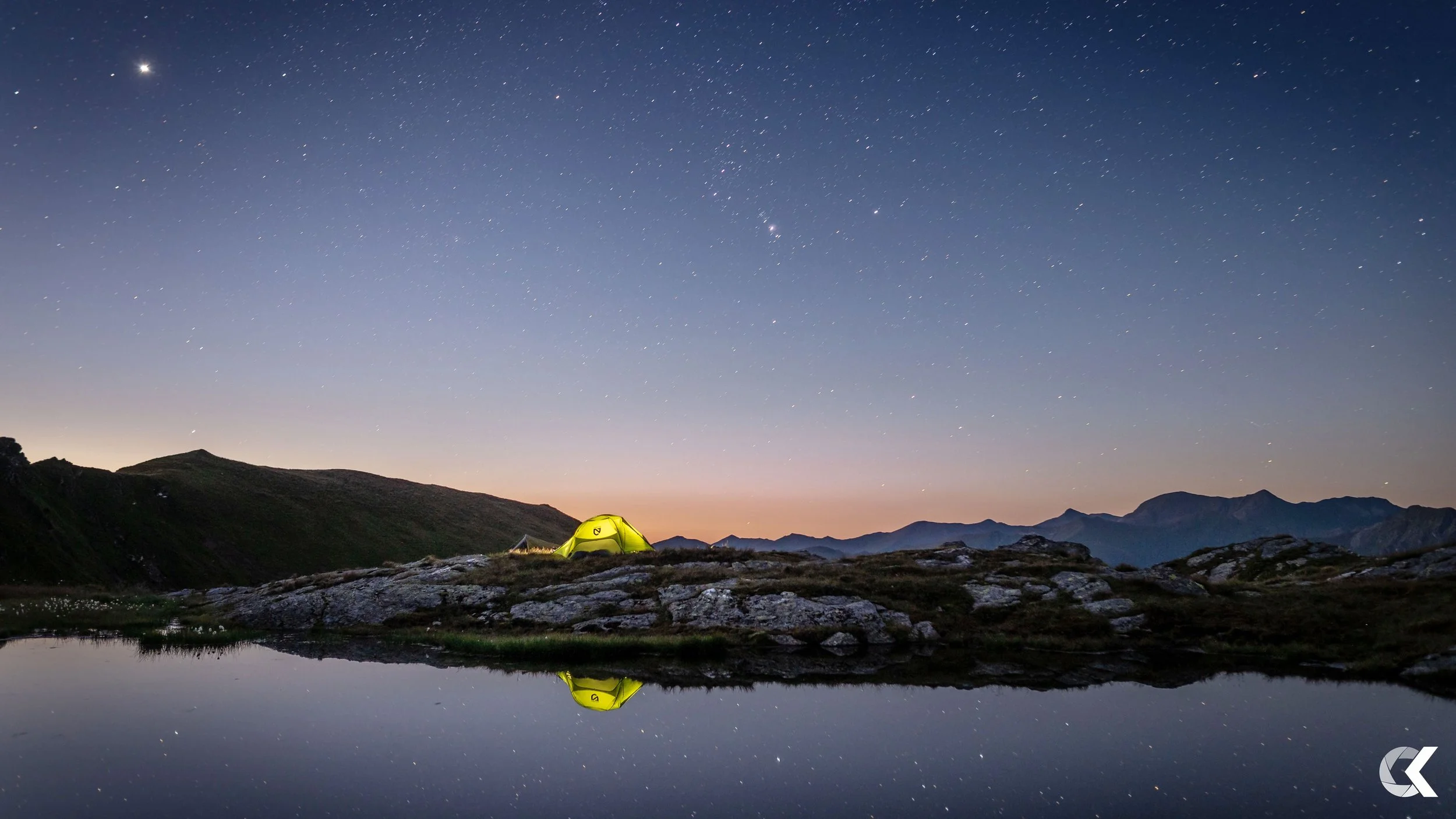 A yellow camping tent set up on rocky terrain by a calm lake at night, with a star-filled sky and mountain silhouettes in the background, reflecting in the lake.