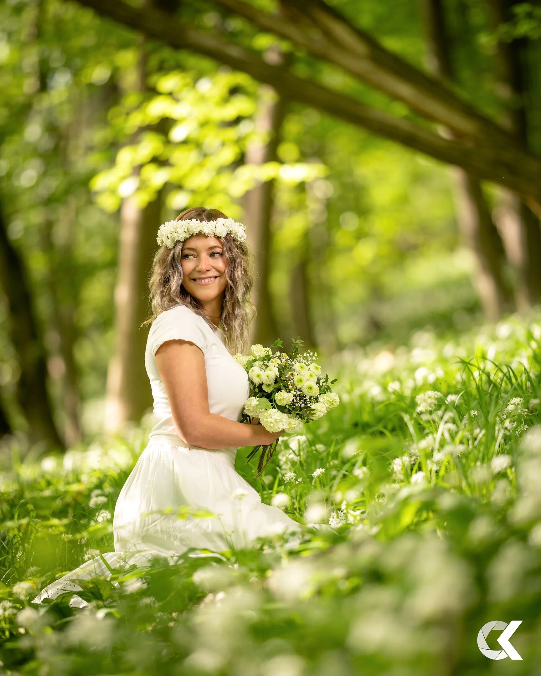 A woman in a white dress with curly hair, wearing a flower crown, is sitting in a green forest holding a bouquet of white flowers, smiling and looking to her left.