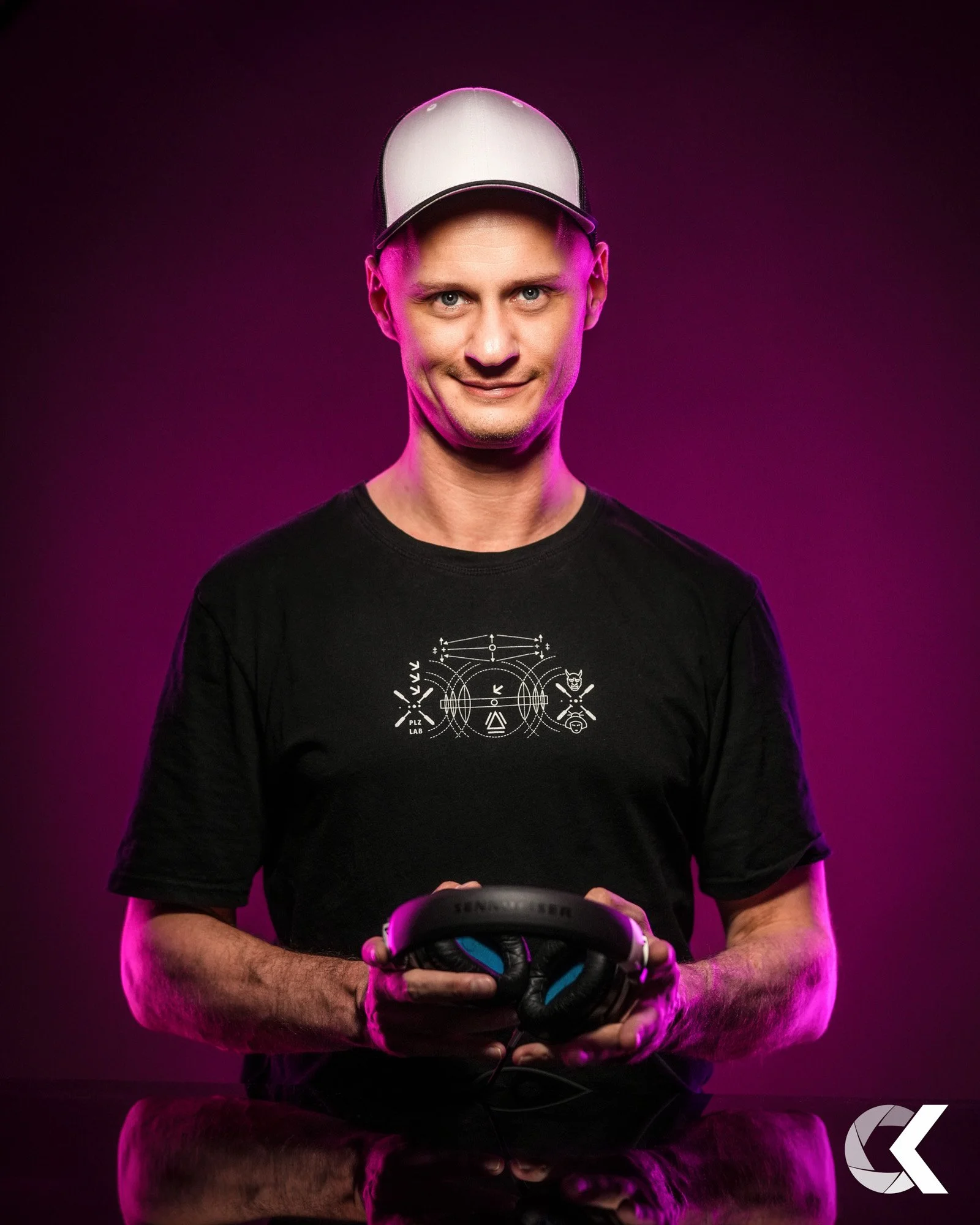 Ein junger Mann trägt eine weiße Kappe, ein schwarzes T-Shirt mit geometrischen Linien und Symbolen, und hält Kopfhörer in den Händen. Er steht vor einem dunklen Hintergrund mit lila Beleuchtung.