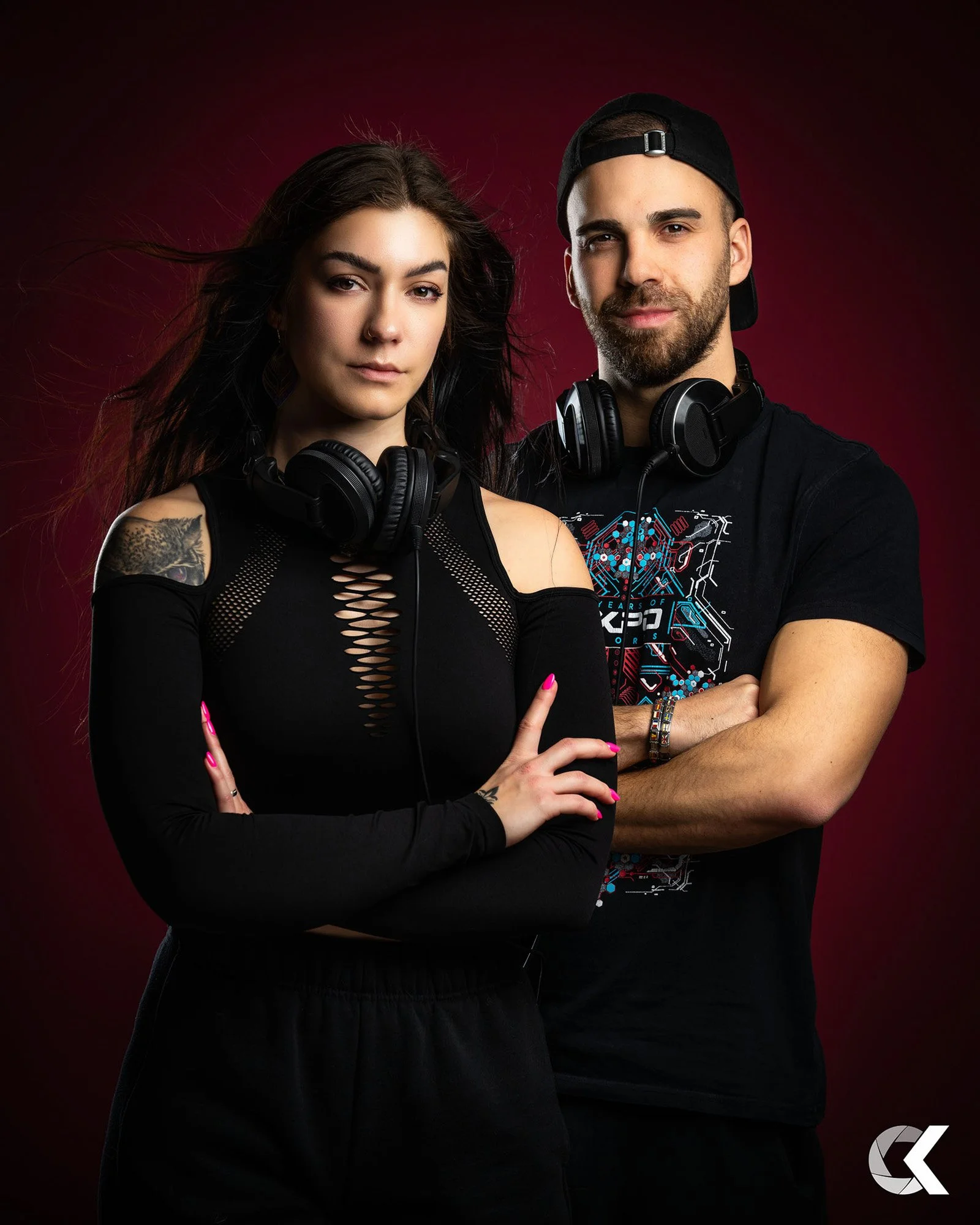 A young woman and man stand side by side against a dark red background, both with headphones around their necks, confident expressions, and arms crossed.