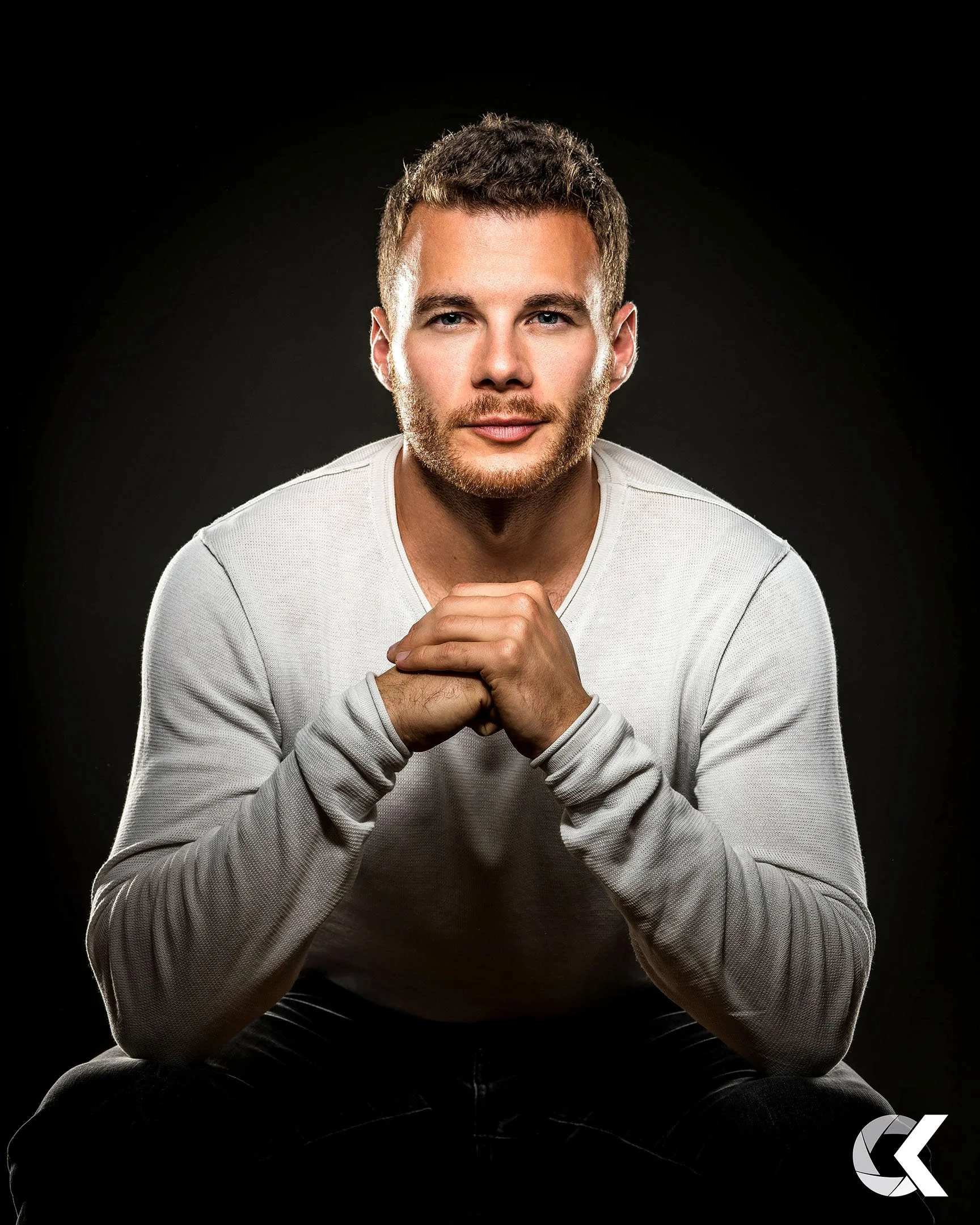 A young man with short brown hair and a beard, wearing a white long sleeve shirt, sitting with his hands clasped and looking into the camera against a dark background.