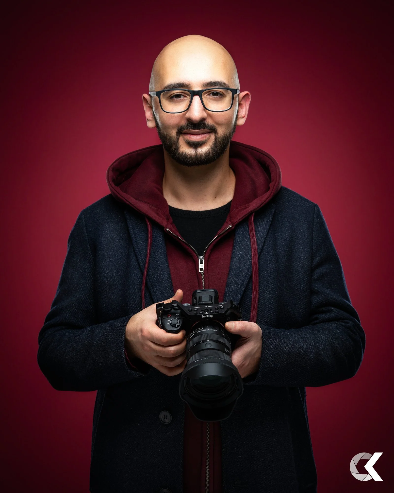 A man with glasses, a beard, and a bald head, holding a professional camera, dressed in a dark jacket with a burgundy hoodie underneath, standing against a dark red background.