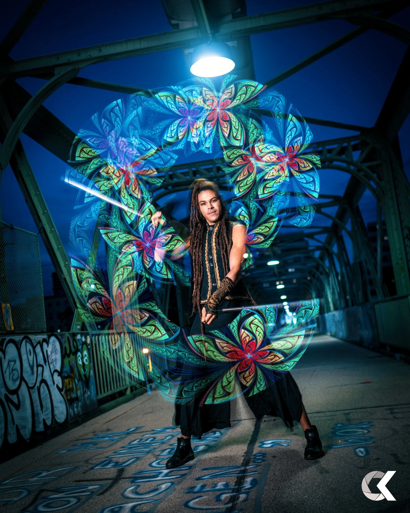 A person with long dreadlocks wearing a black and gold outfit, standing on a graffiti-covered sidewalk under a bridge at night, performing with illuminated light painting creating swirling colorful floral patterns around them.