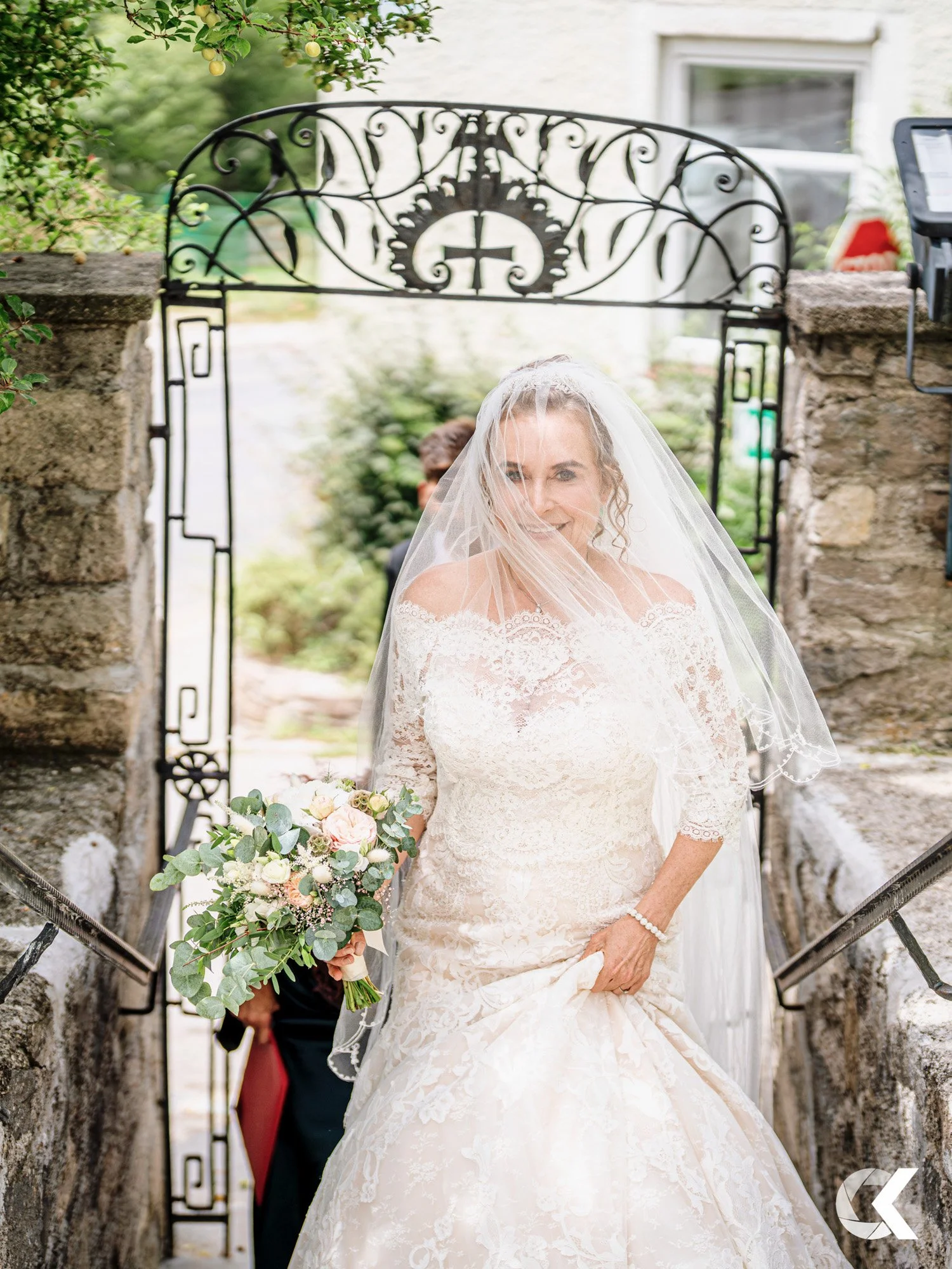 A bride in a lace wedding dress and veil, holding a bouquet of flowers, is smiling and walking through an iron gate with stone walls on either side, on a garden path.