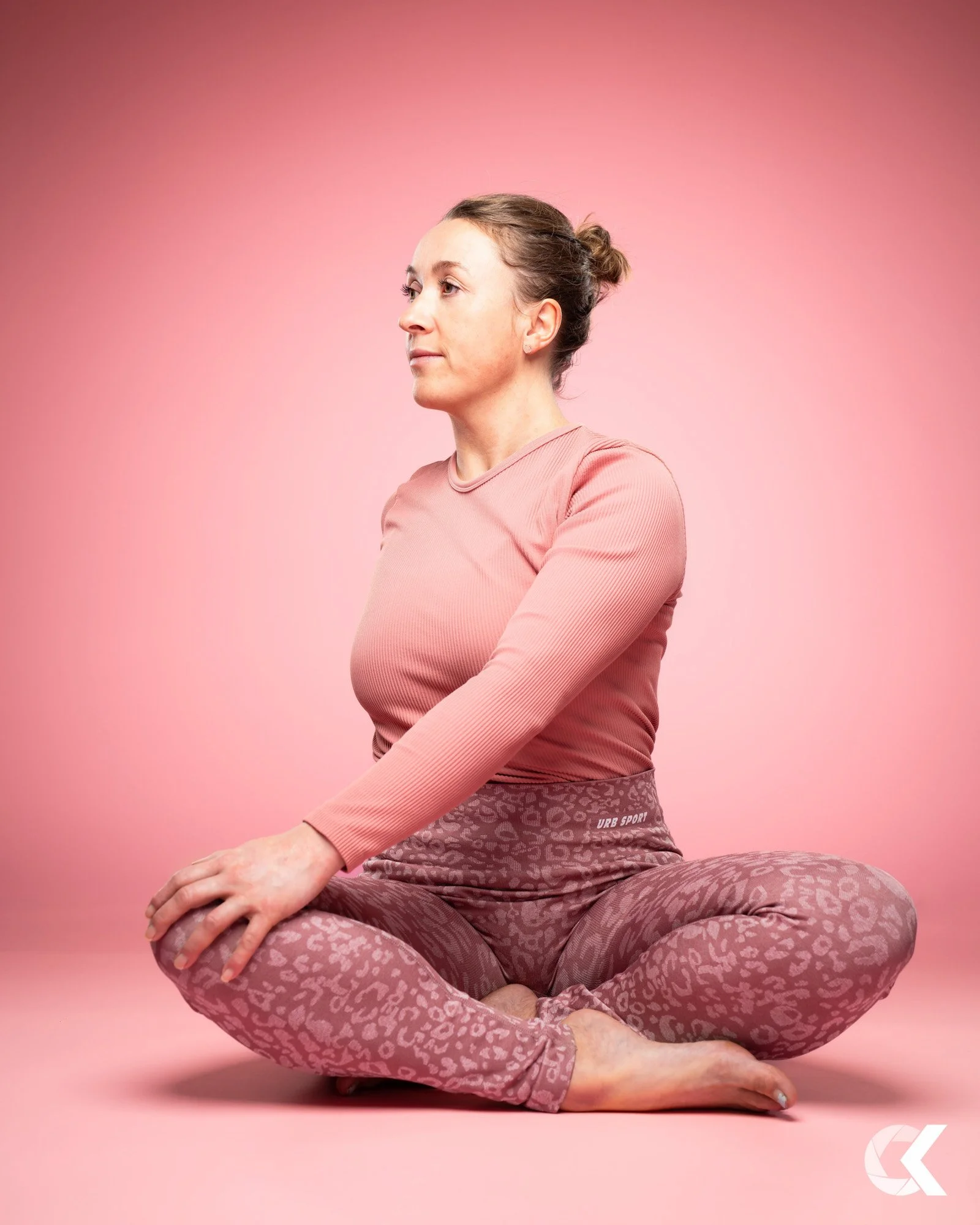 Frau in Yoga-Pose, sitzend auf dem Boden gegen einen pinken Hintergrund.