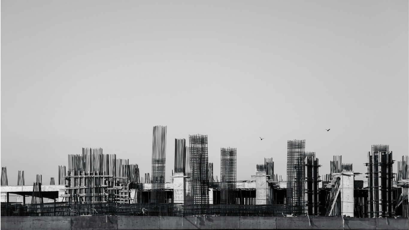 Black and white photo of a construction site with multiple concrete columns and steel reinforcement bars, some partially built, against a plain gray sky.