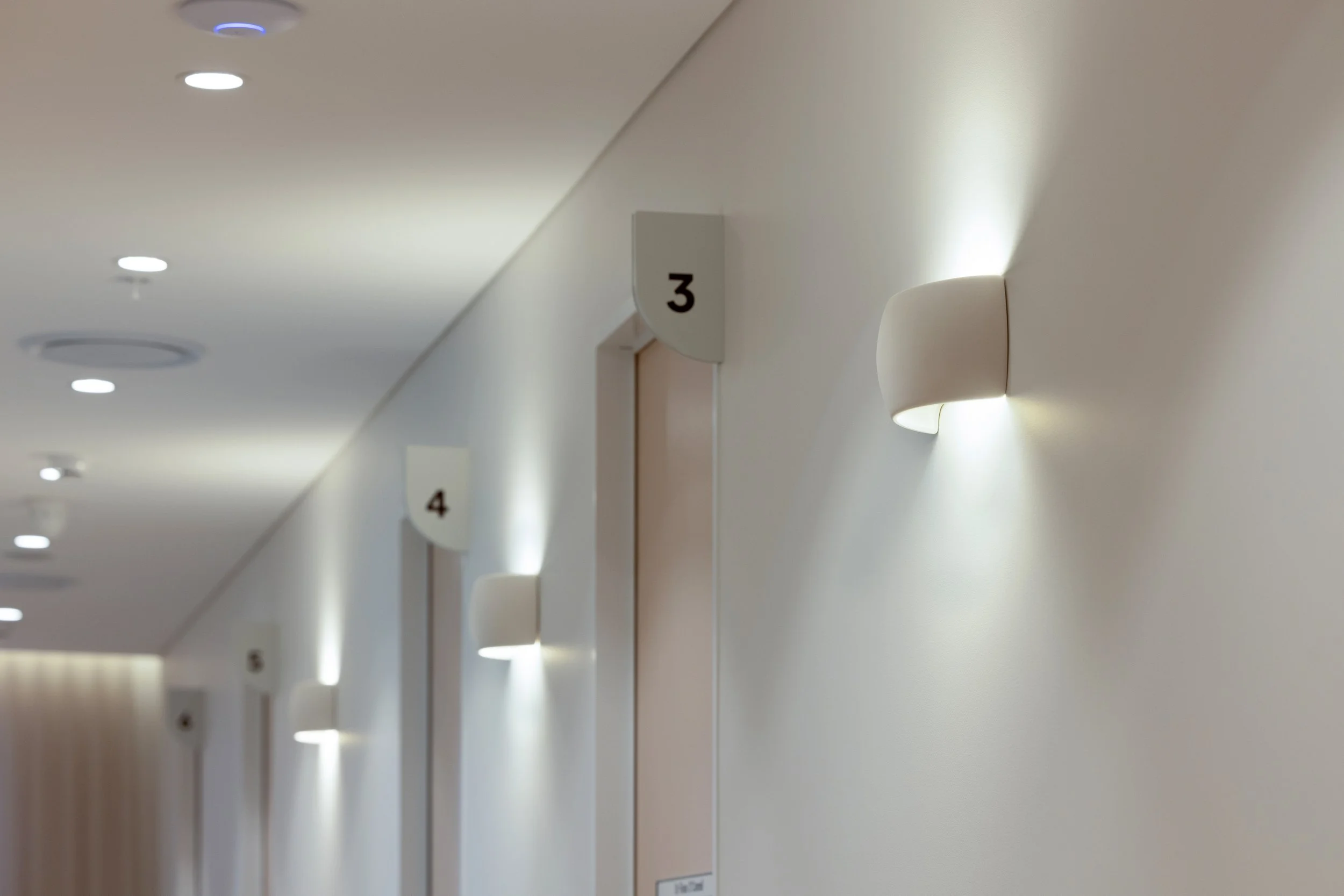 Hospital hallway with evenly spaced wall-mounted lights, numbered signs on the wall indicating different room numbers, with room 3 visible in the foreground and room 4 further down the corridor.