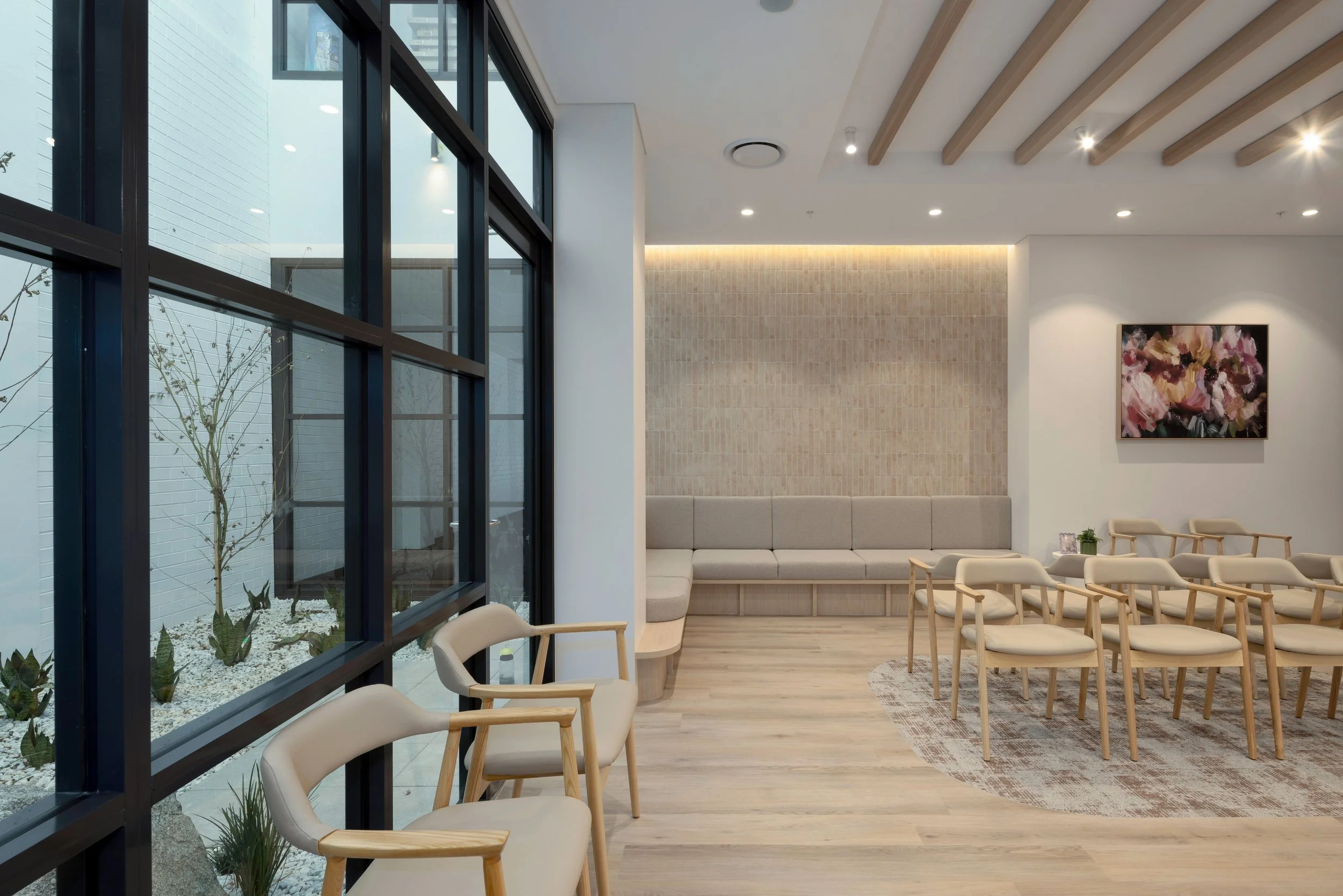 Modern interior space with seating area, large abstract flower painting, and a view of outdoor greenery through large glass windows.
