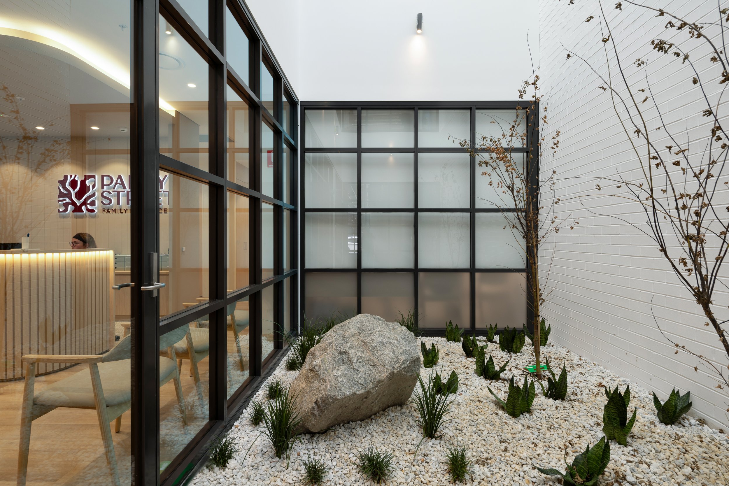 Indoor courtyard with white gravel, a large rock, small plants, and a white brick wall with leafless branches. Modern office space visible through glass panels with a reception desk and a woman working.
