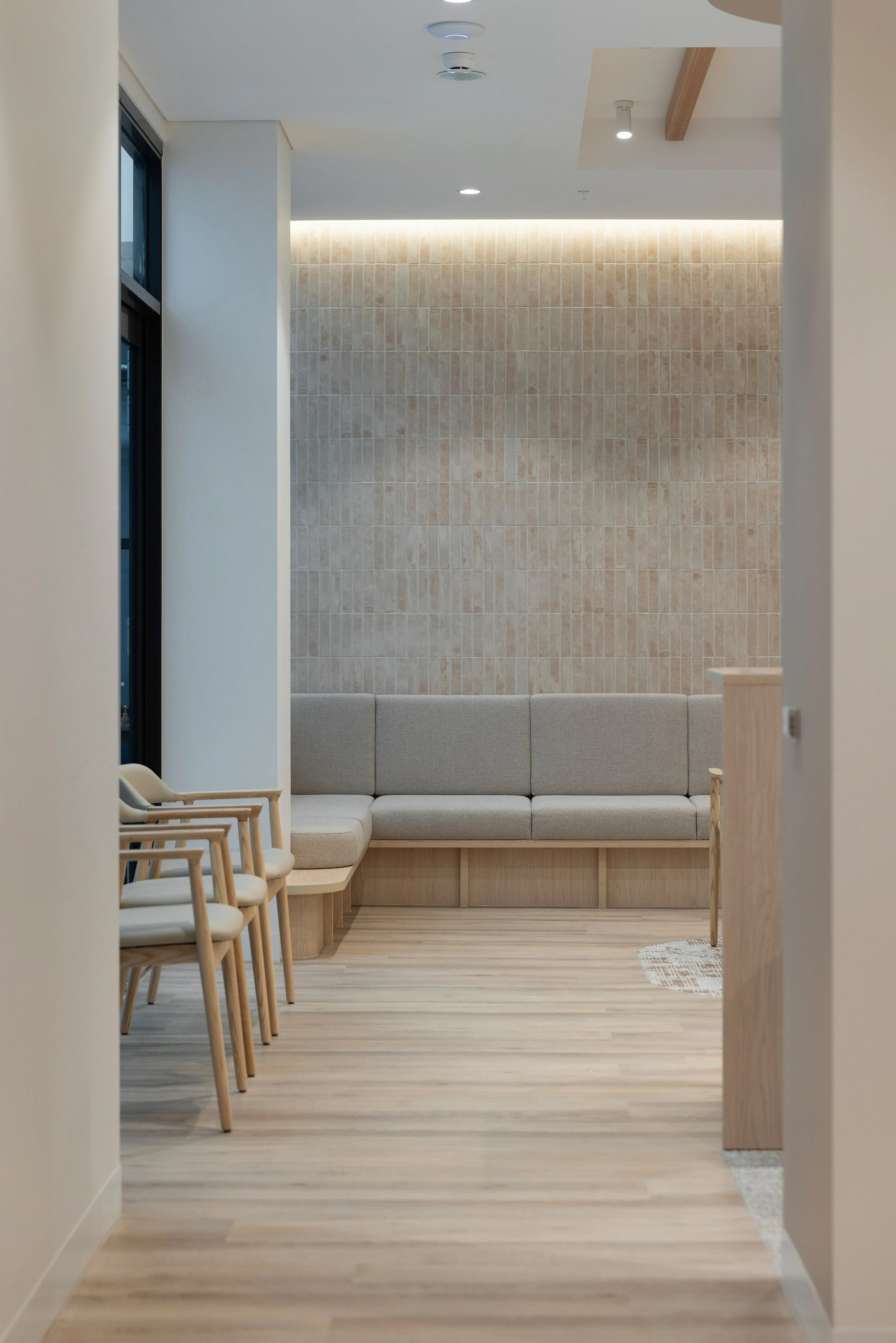 A waiting area with beige chairs, a long beige bench seat, light wood flooring, a beige tiled wall, and natural lighting.