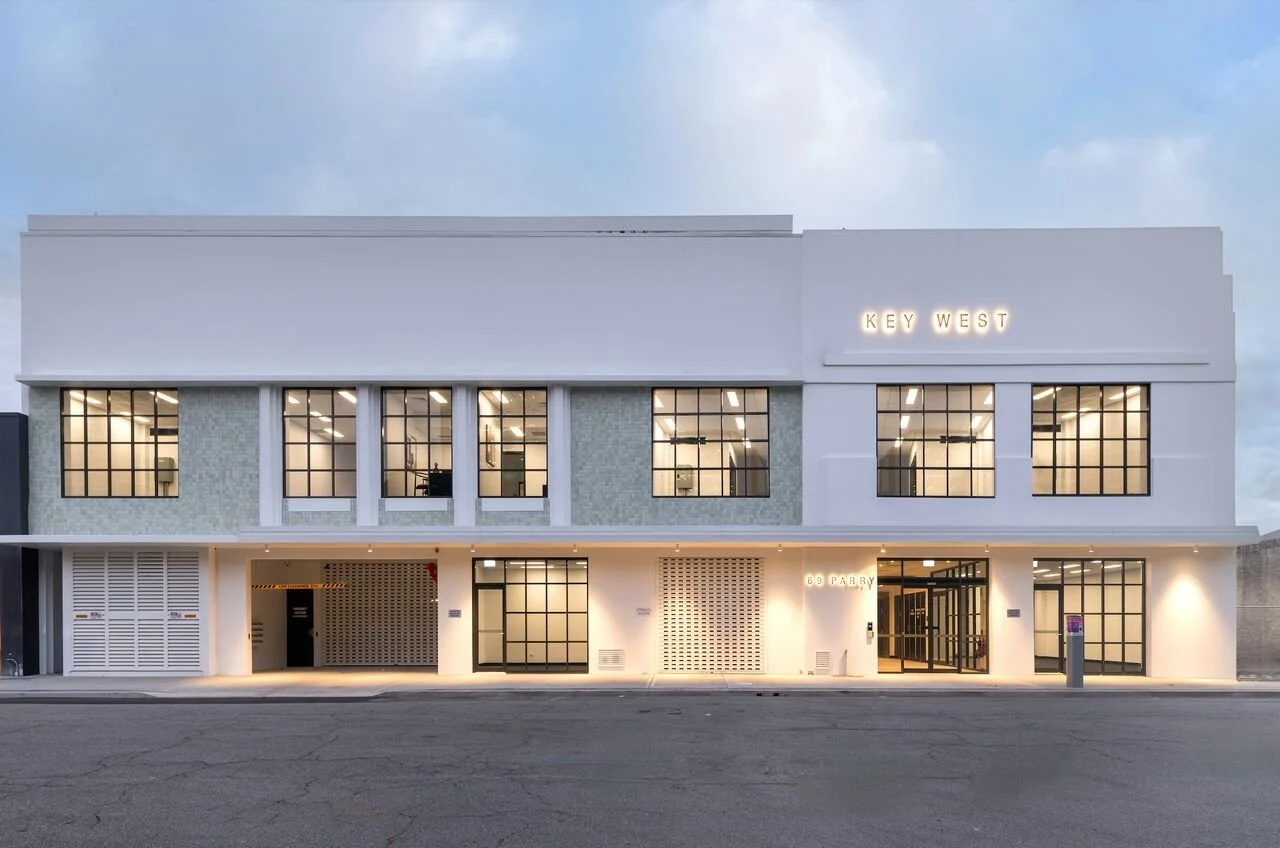 Modern two-story building with large glass windows, illuminated interior, and neon sign reading "KEY WEST" on the upper right.