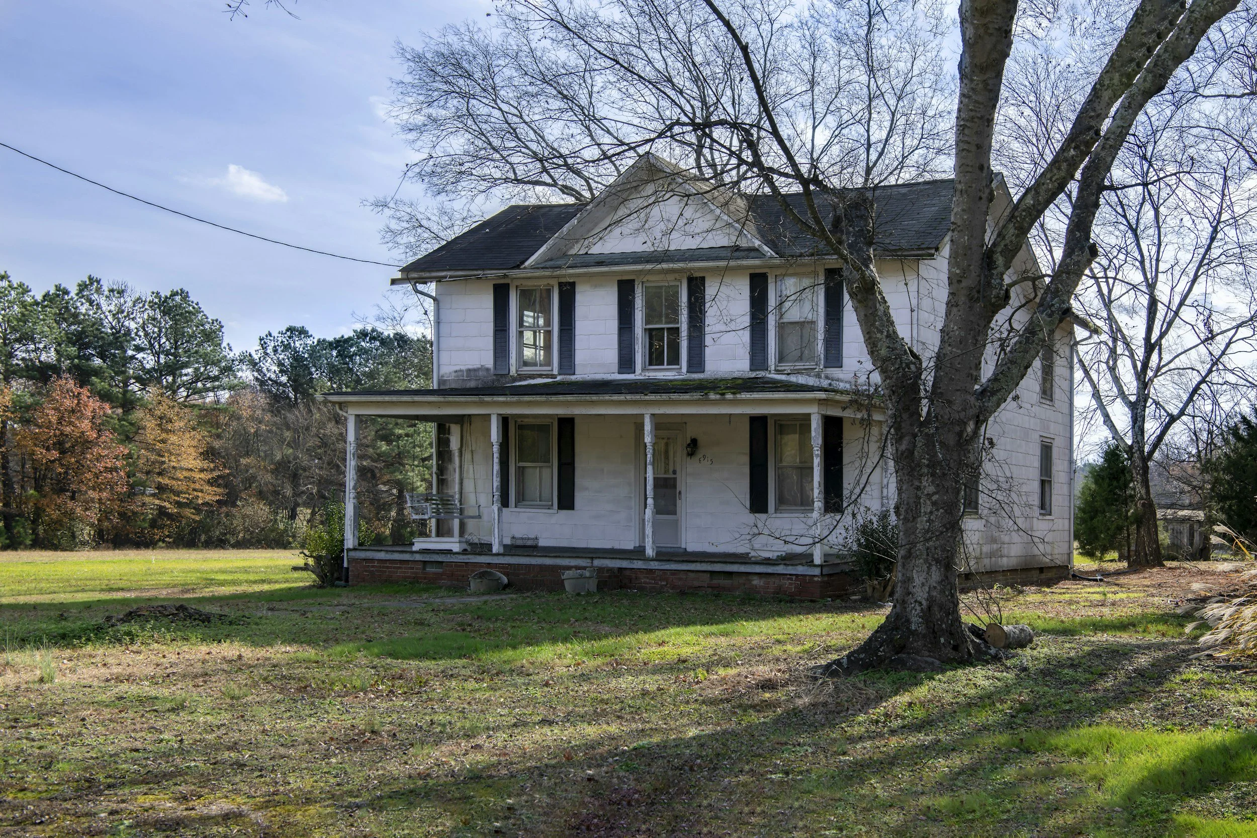 An old, two-story white house with black shutters, front porch, and a gabled roof surrounded by trees, some of which have lost their leaves, on a grass yard under a partly cloudy sky.