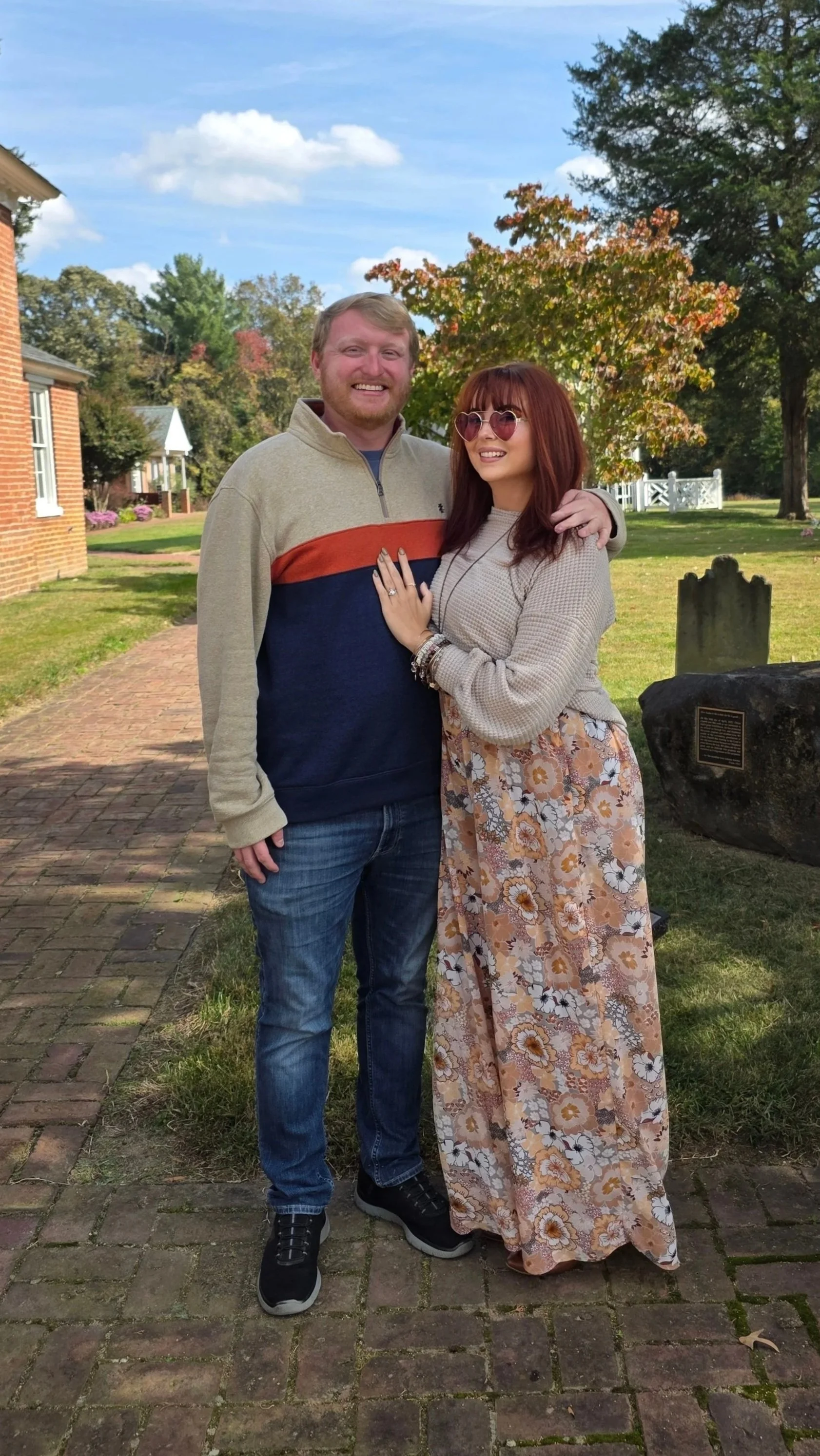 A couple standing outdoors on a brick walkway, smiling. The woman is wearing pink heart-shaped sunglasses, a beige sweater, and a long floral skirt. The man is wearing a beige and navy sweater with a red stripe, jeans, and black sneakers. They are surrounded by trees with green and autumn-colored leaves, with houses and a plaque on a large rock visible in the background.