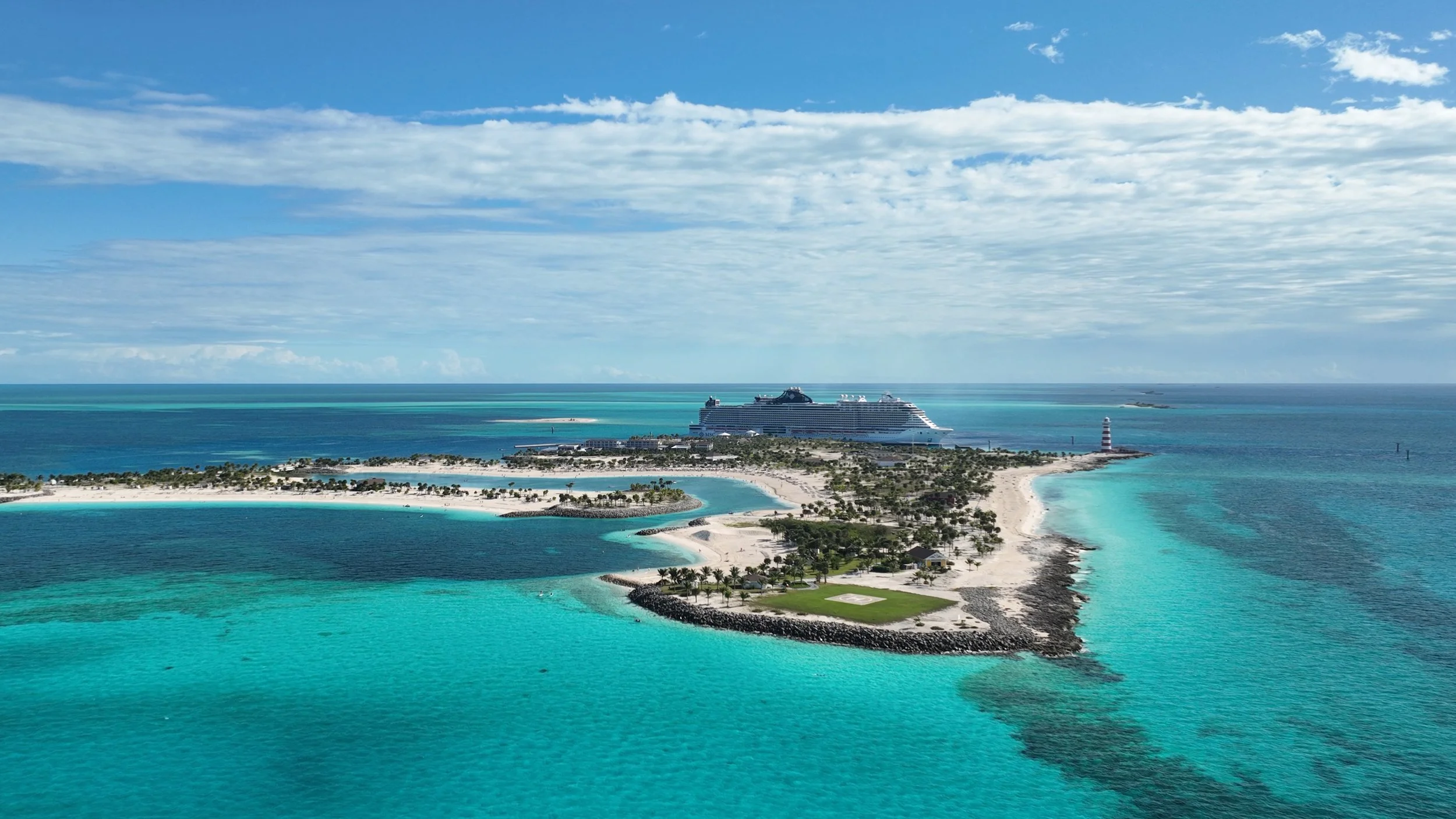 Aerial view of a tropical island with white sandy beaches, clear turquoise waters, palm trees, and a cruise ship in the distance, under a partly cloudy sky.