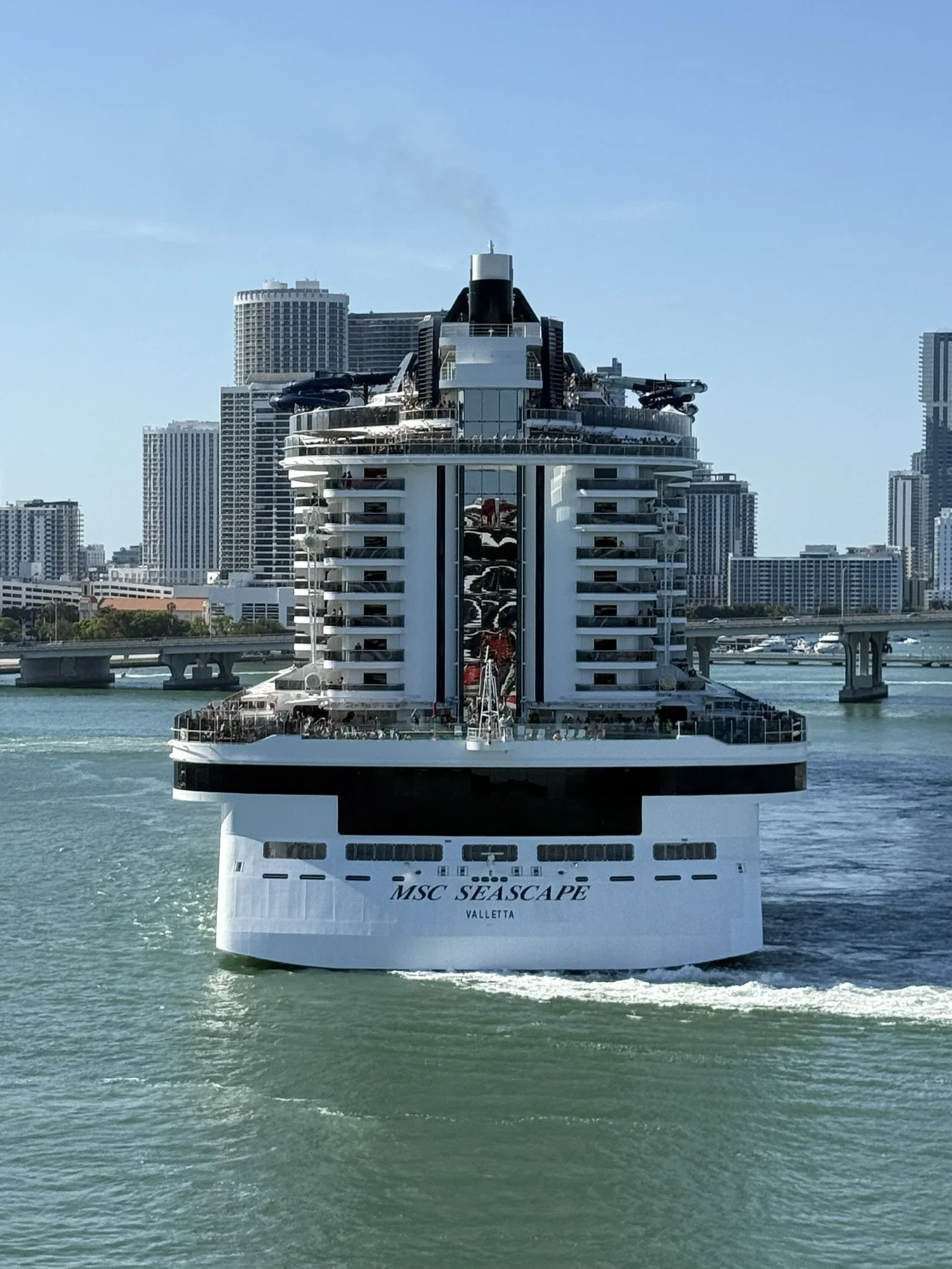 Luxury cruise ship MSC Seascape sailing through a city harbor with skyscrapers in the background.