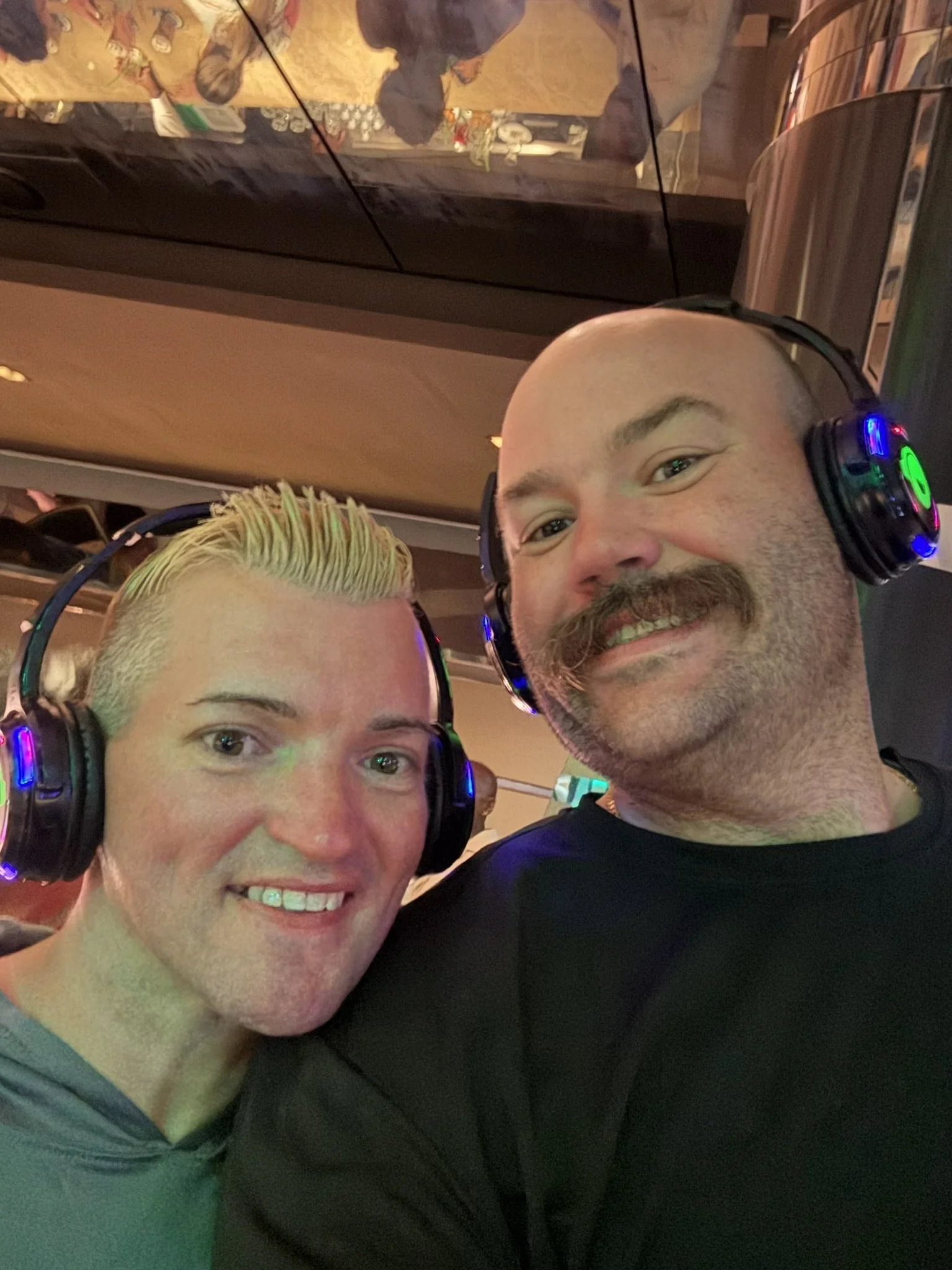 Two men wearing headphones smiling at the camera, with a mirror reflecting their image and a curved metallic surface in the background.